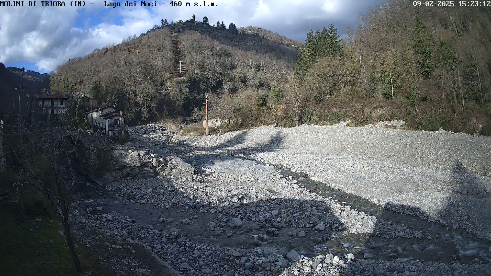 A rocky, shallow riverbed with a small stream of water flows through it, nestled between buildings and a hillside covered in leafless trees under partly cloudy skies.