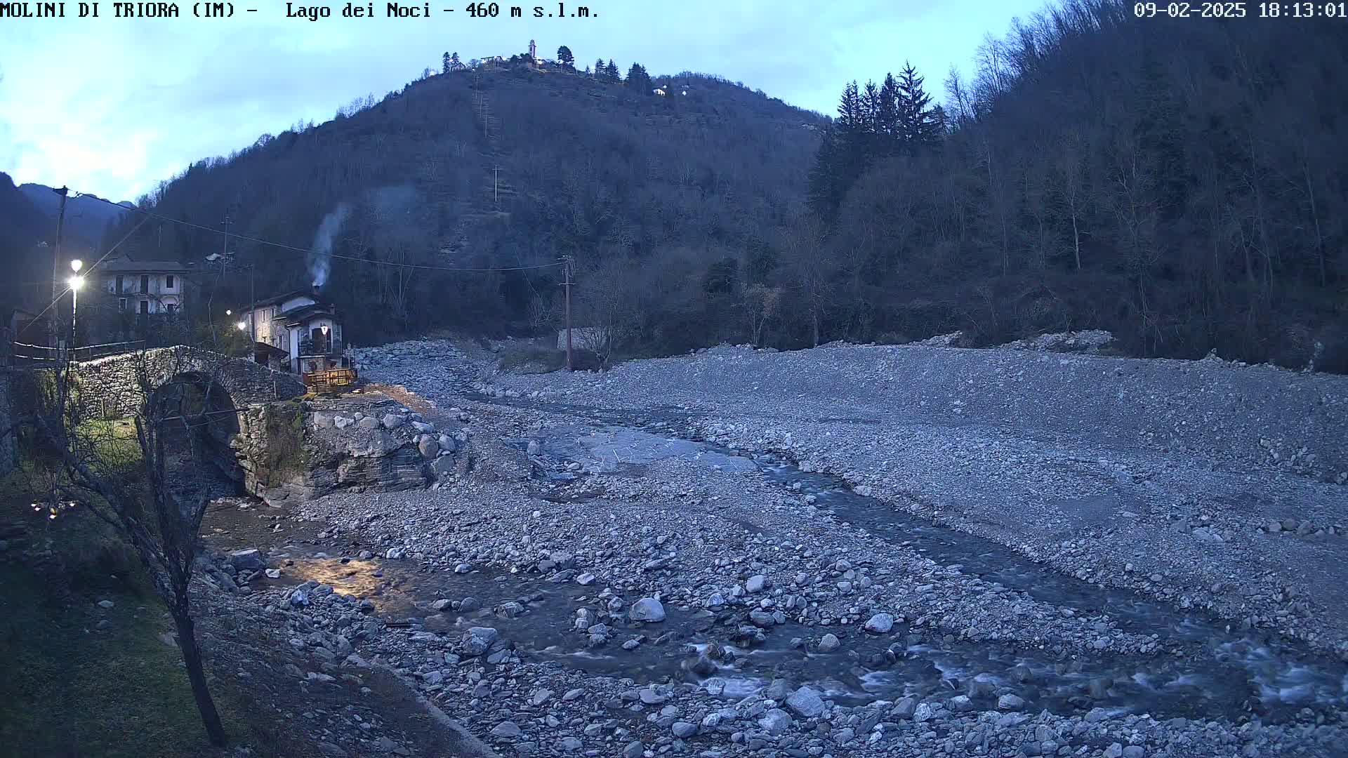 A stone bridge crosses a shallow, rocky stream under a twilight sky, with a village and hill visible in the background.