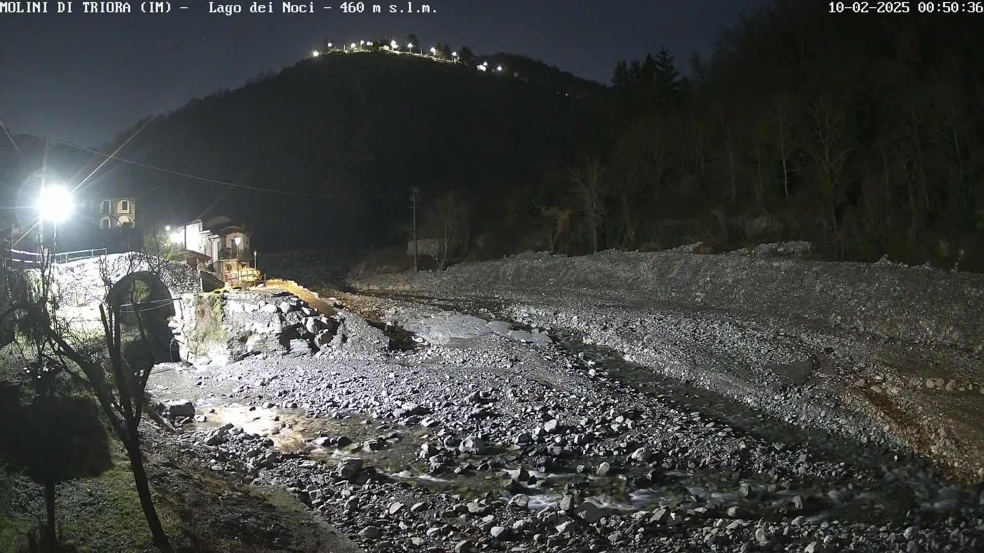 A rocky, shallow stream flows under a stone arch bridge at night, illuminated by artificial lights, with a dark hill in the background.
