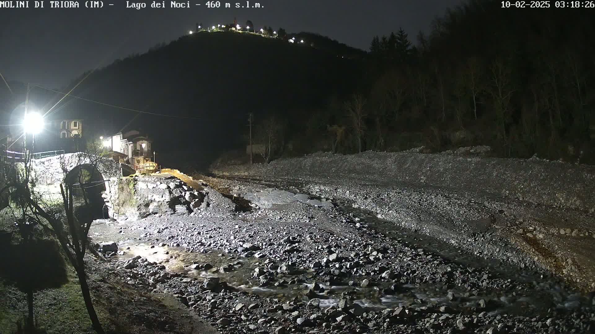 A nighttime view of a rocky riverbed with a stone bridge and buildings illuminated on a hill in the background under a dark sky.