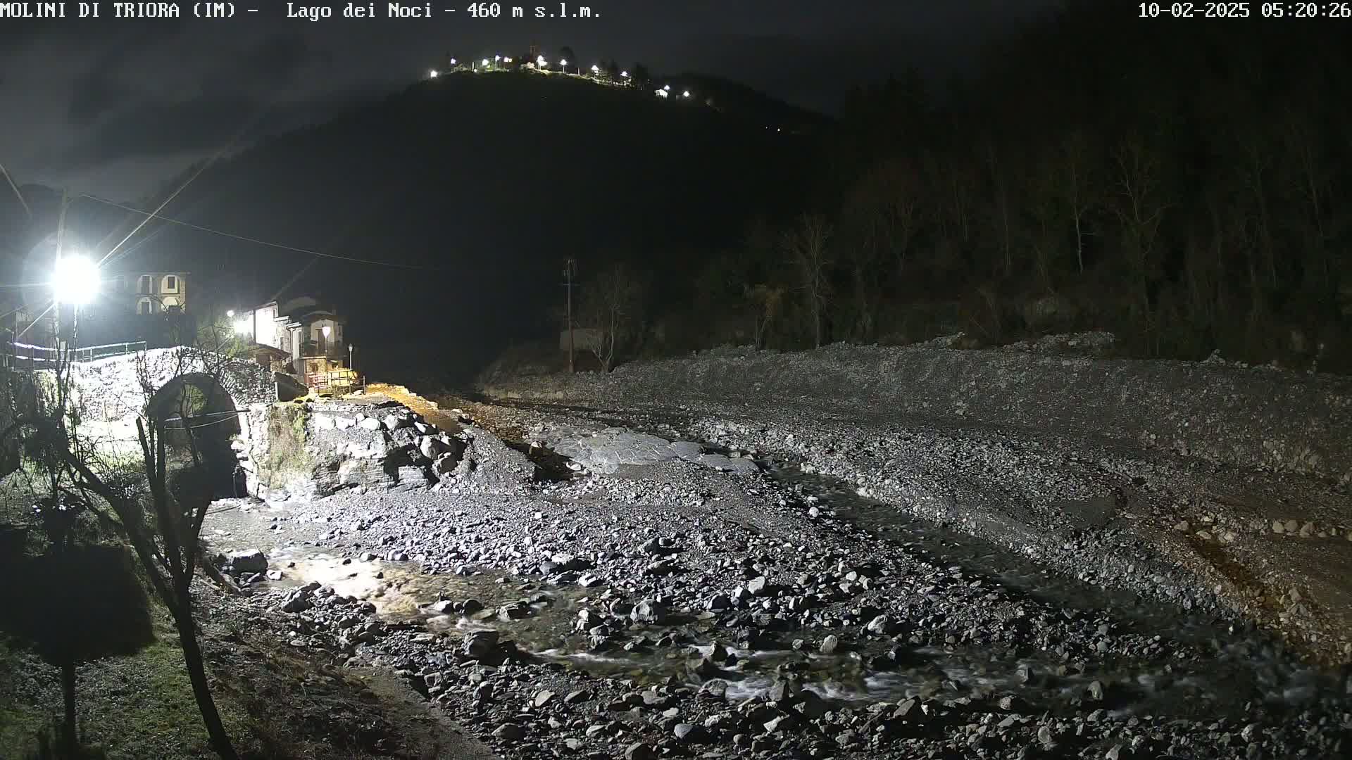 A nighttime view of a rocky stream flowing under a stone arch bridge, with buildings and hills in the background under dark skies.