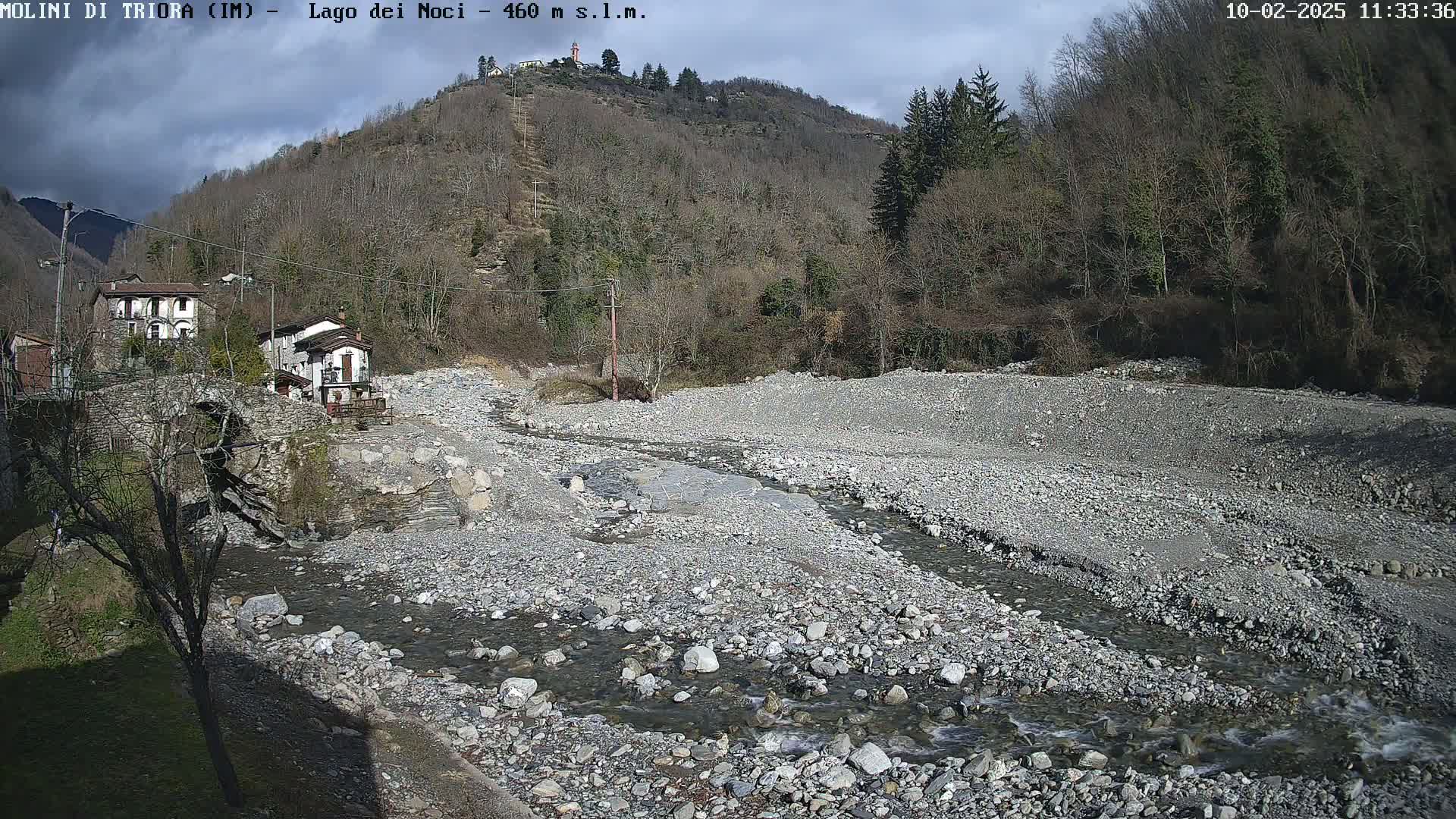 A rocky, mostly dry riverbed with a small stream running through it, situated below a village nestled against a hillside of leafless trees under a partly cloudy sky.