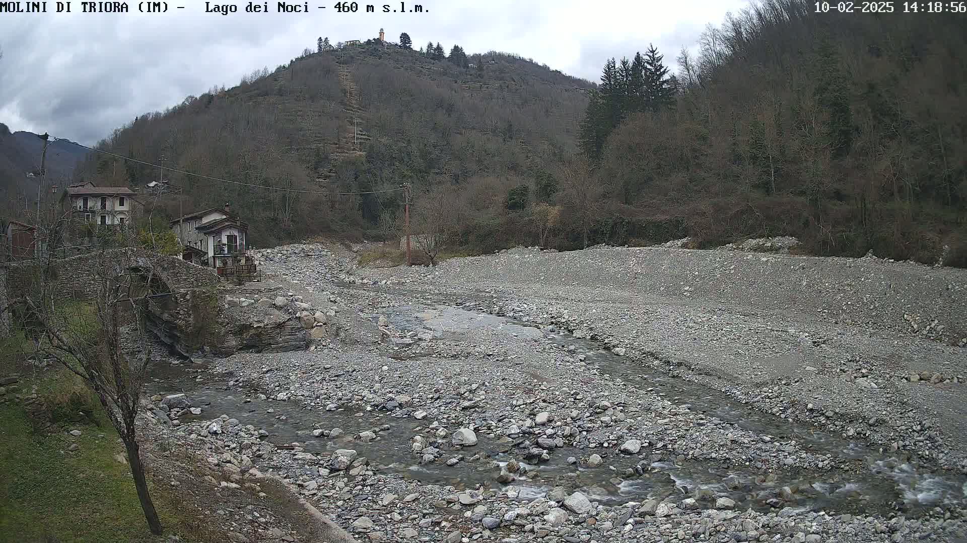A rocky, mostly dry riverbed with a small stream flows through it, past a stone bridge and houses nestled against a hillside under an overcast sky.