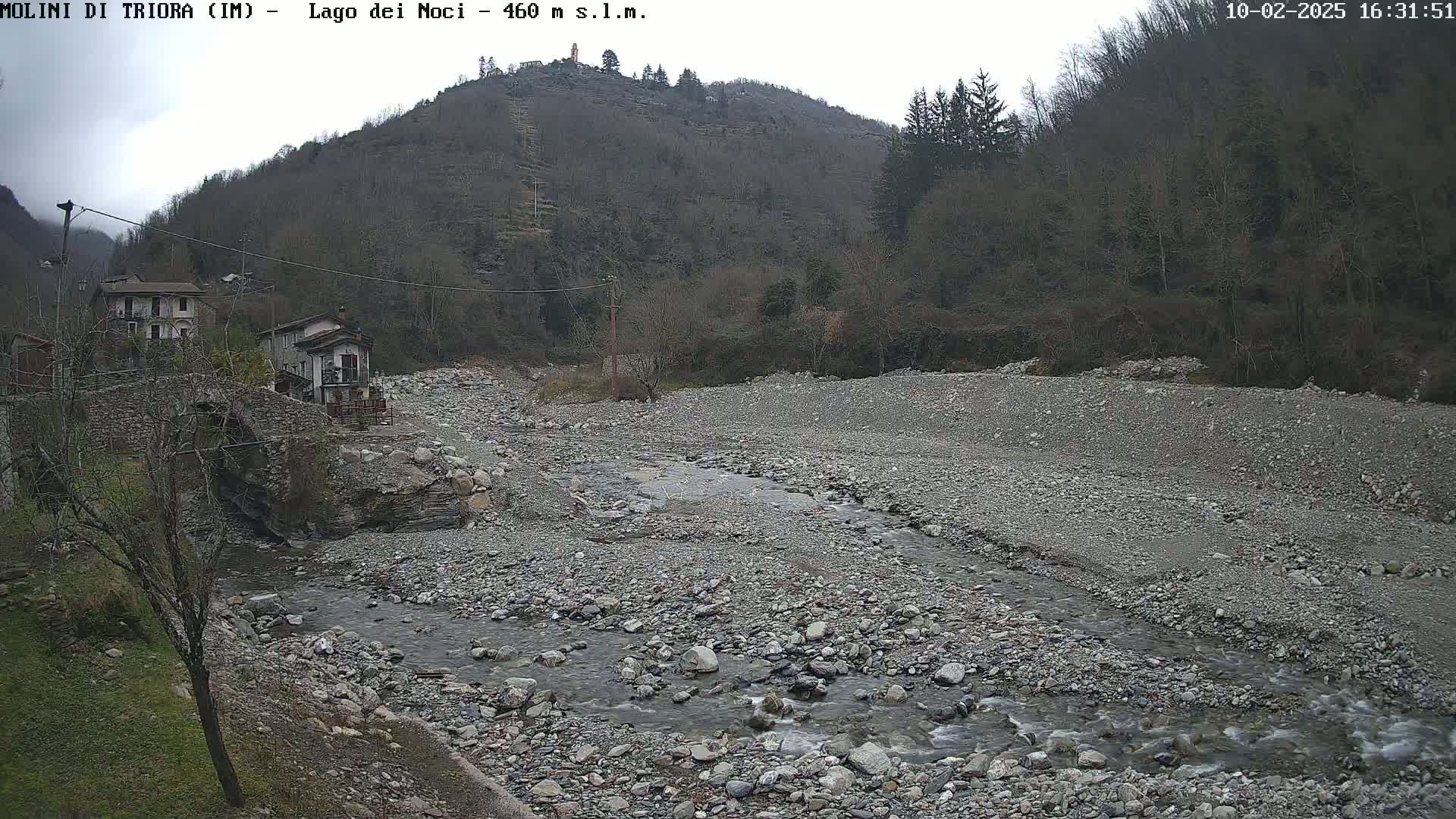 A rocky, shallow river flows through a sparsely vegetated valley beneath an overcast sky, past a stone bridge and a few buildings nestled against a hillside.