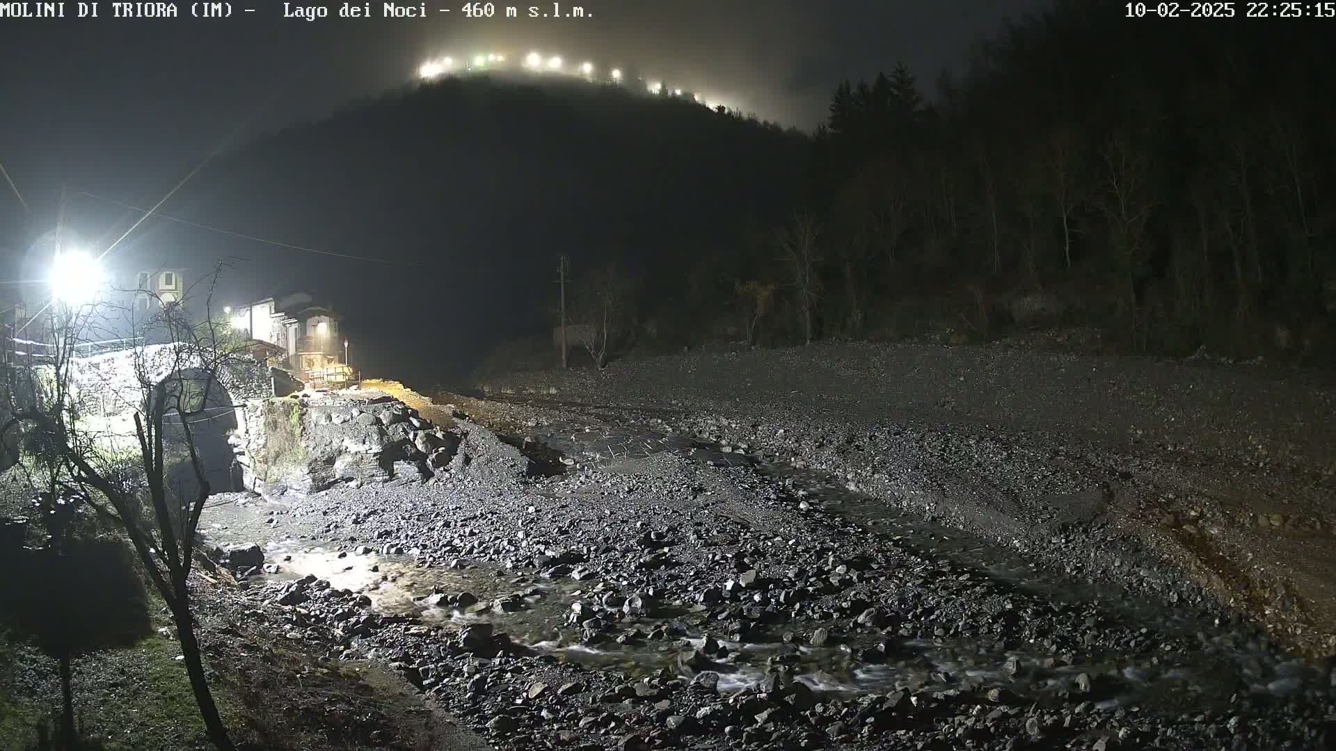 A nighttime scene shows a rocky streambed with a slow-moving stream under a foggy sky, near a stone bridge and buildings illuminated by artificial lights.