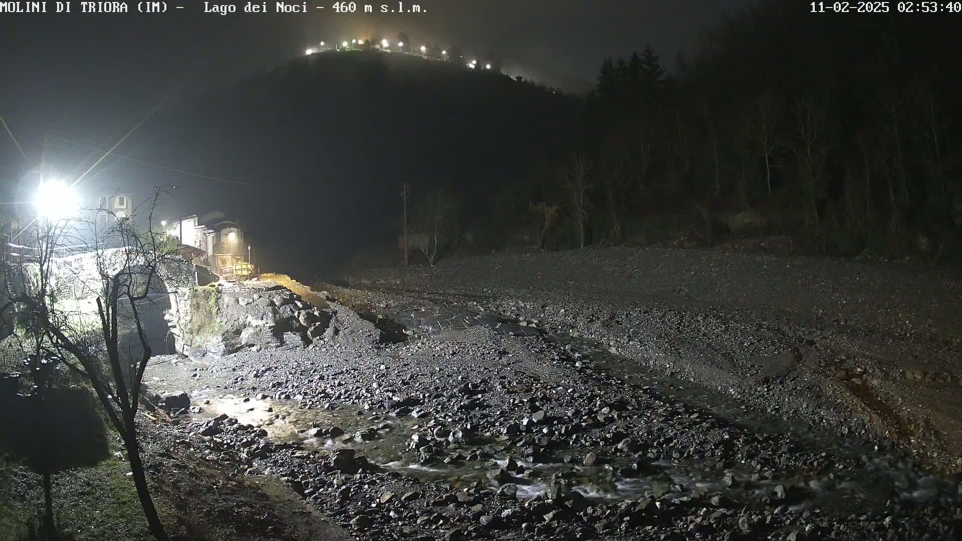A nighttime, low-light view shows a rocky stream flowing under a stone bridge near a village nestled below a hill.