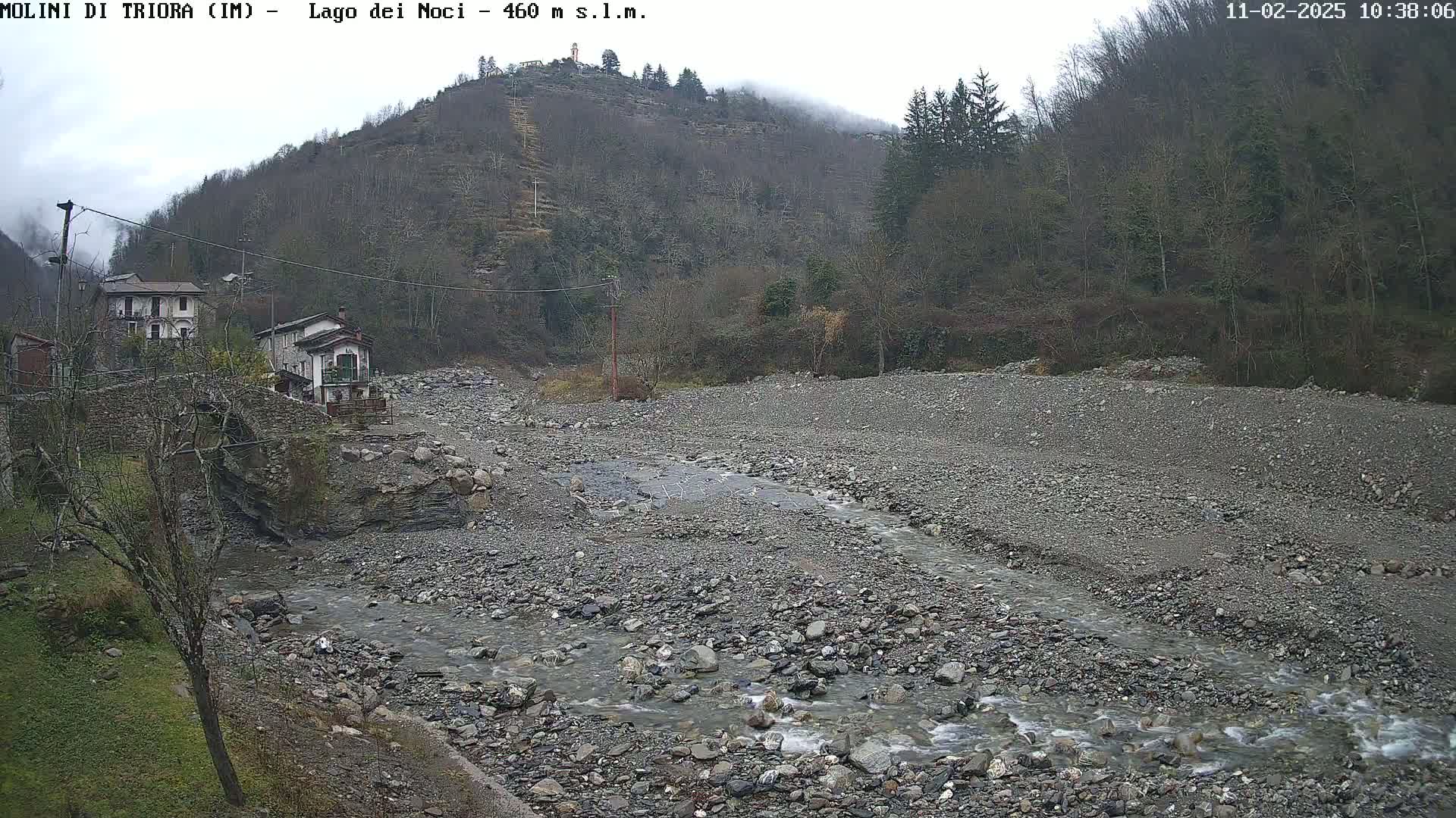 A rocky, dry riverbed with a small stream flows through it, nestled between a hillside covered in leafless trees and a small village under an overcast sky.