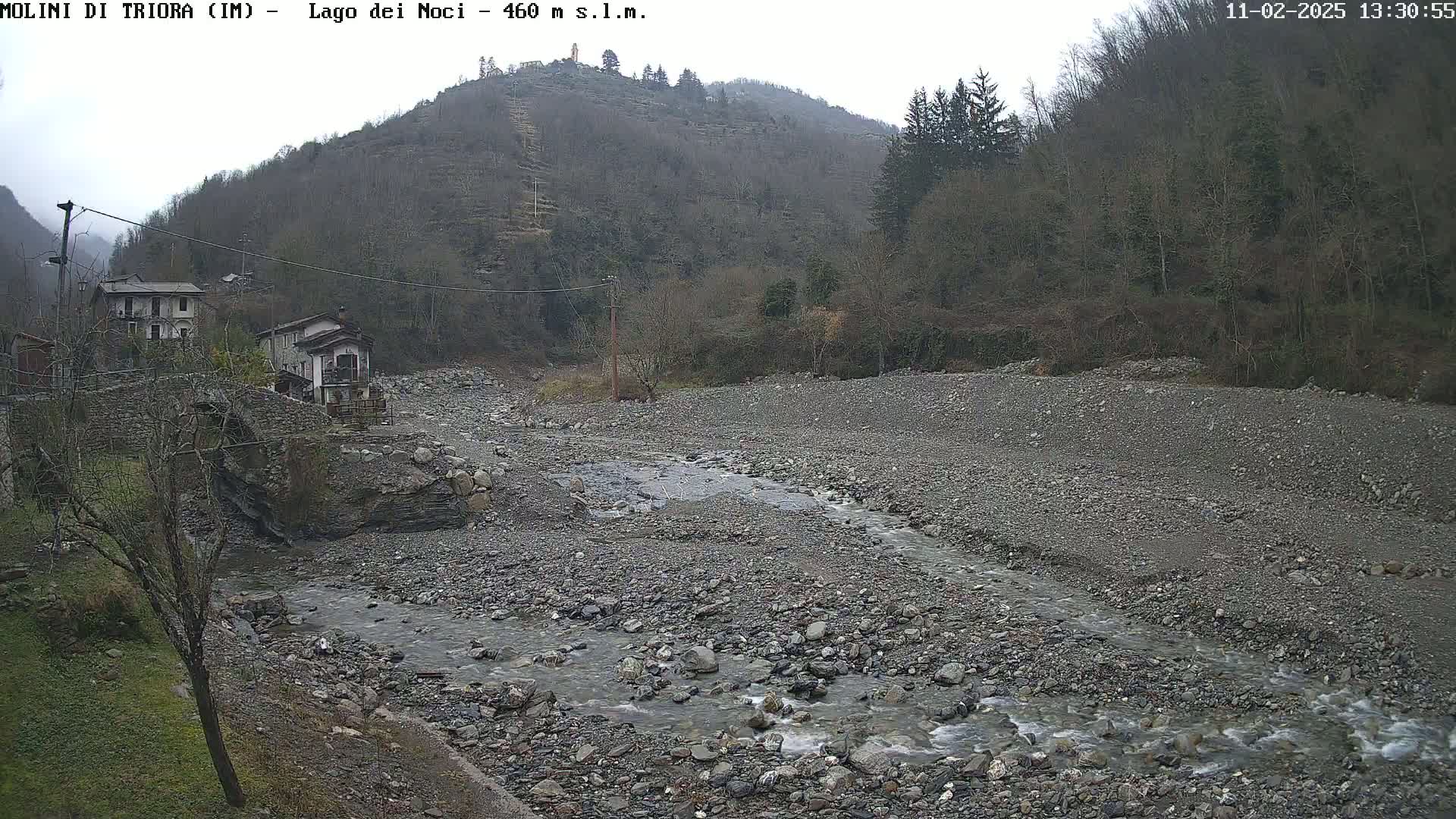 A rocky, shallow stream flows through a dry riverbed, past a small village nestled at the base of a hill covered in leafless trees under an overcast sky.