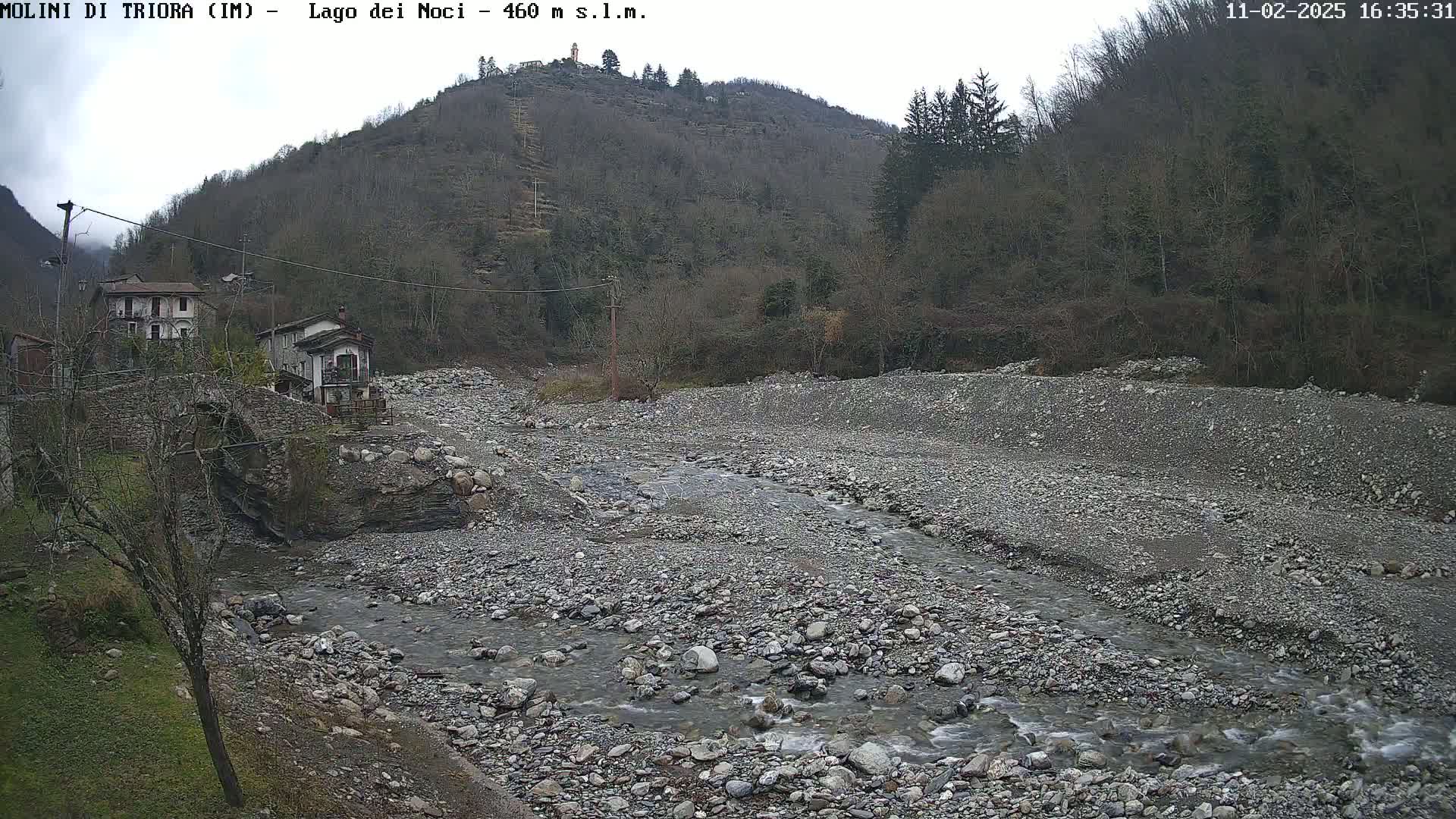 A rocky, shallow stream flows through a dry riverbed, past a stone bridge and small village nestled against a hillside under an overcast sky.