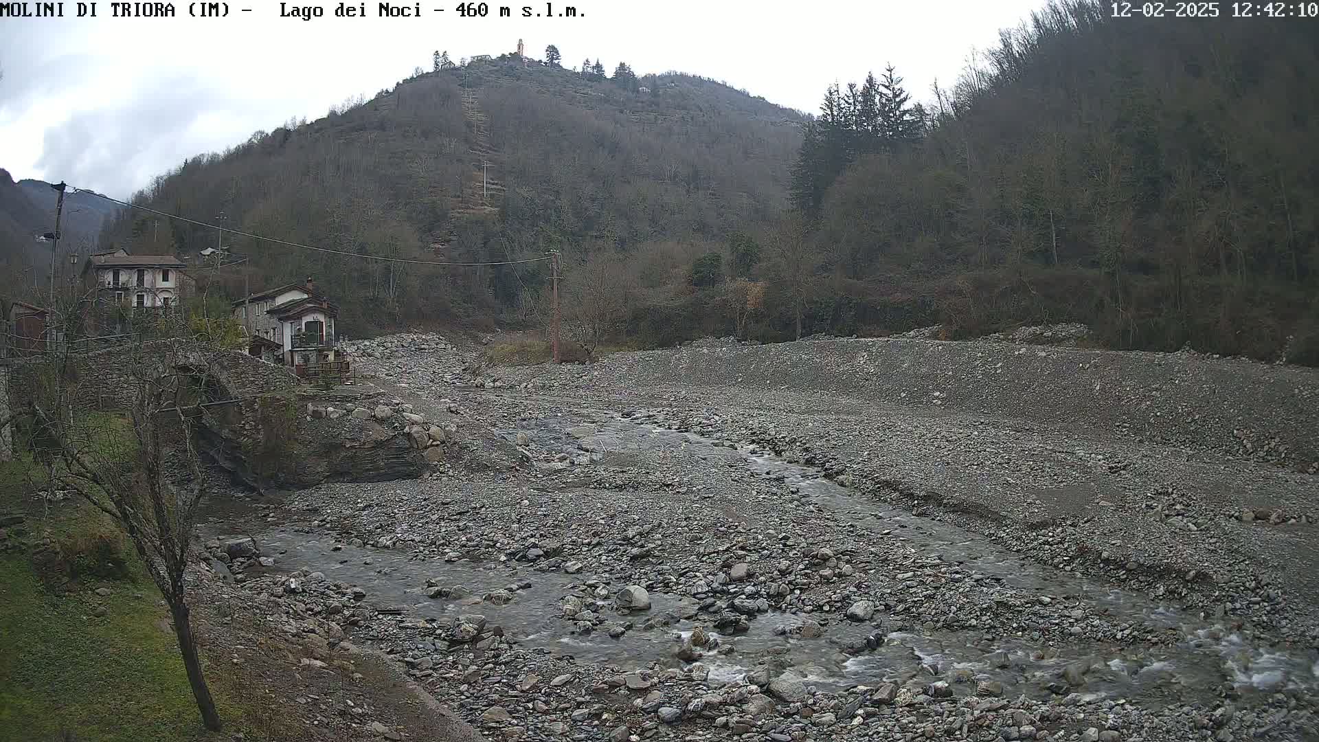 A rocky, dry riverbed with a small stream flows through it, past a stone bridge and houses nestled at the base of a treeless hill under an overcast sky.