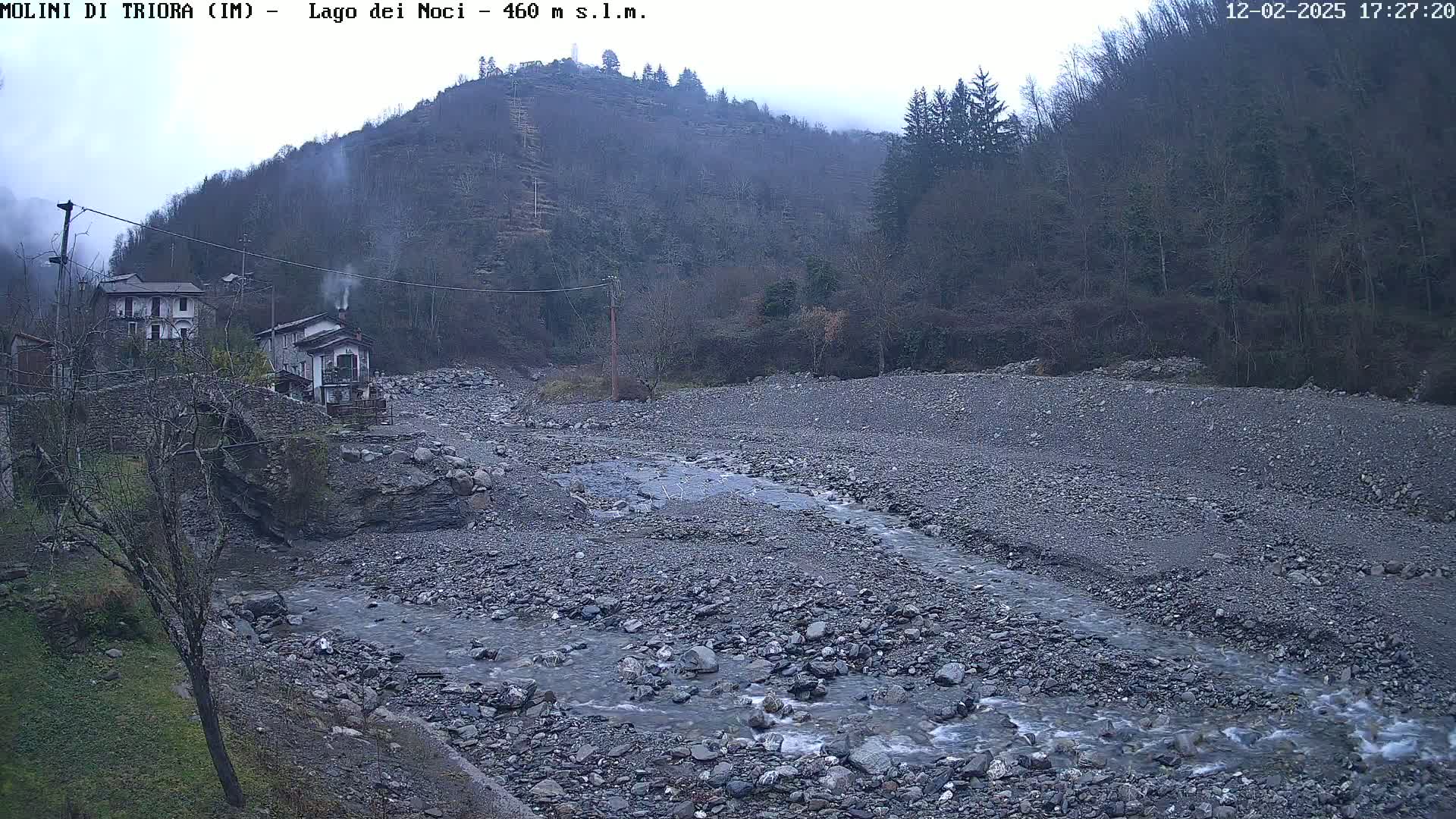 A rocky riverbed with a small stream flows past a village nestled in a valley under an overcast sky.