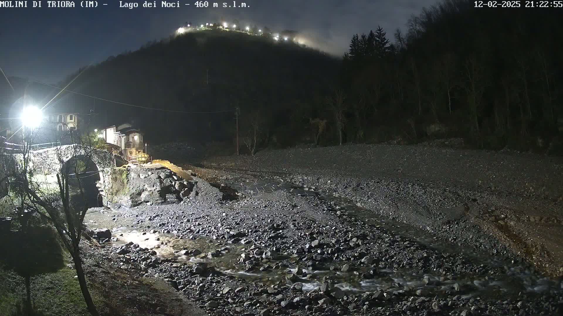 A rocky stream flows under a stone arch bridge at night, with a hilltop illuminated by lights in the background.