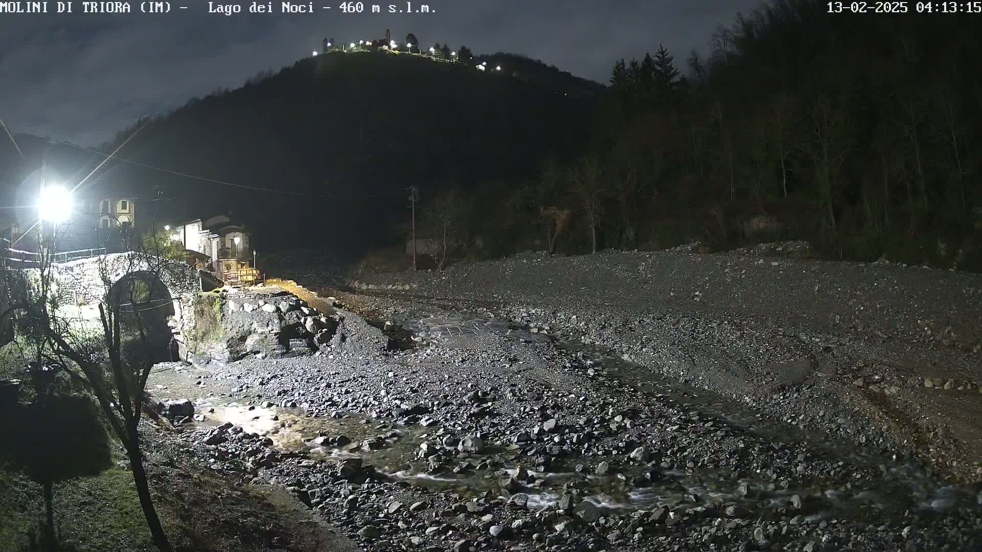 A nighttime view of a rocky stream flowing under a stone bridge near buildings, with a hill in the background under a dark sky.