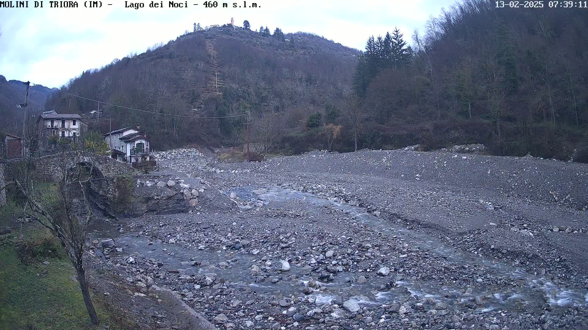 A rocky riverbed with a slow-moving stream winds through a valley under an overcast sky, past a stone bridge and several buildings nestled against a hillside covered in leafless trees.