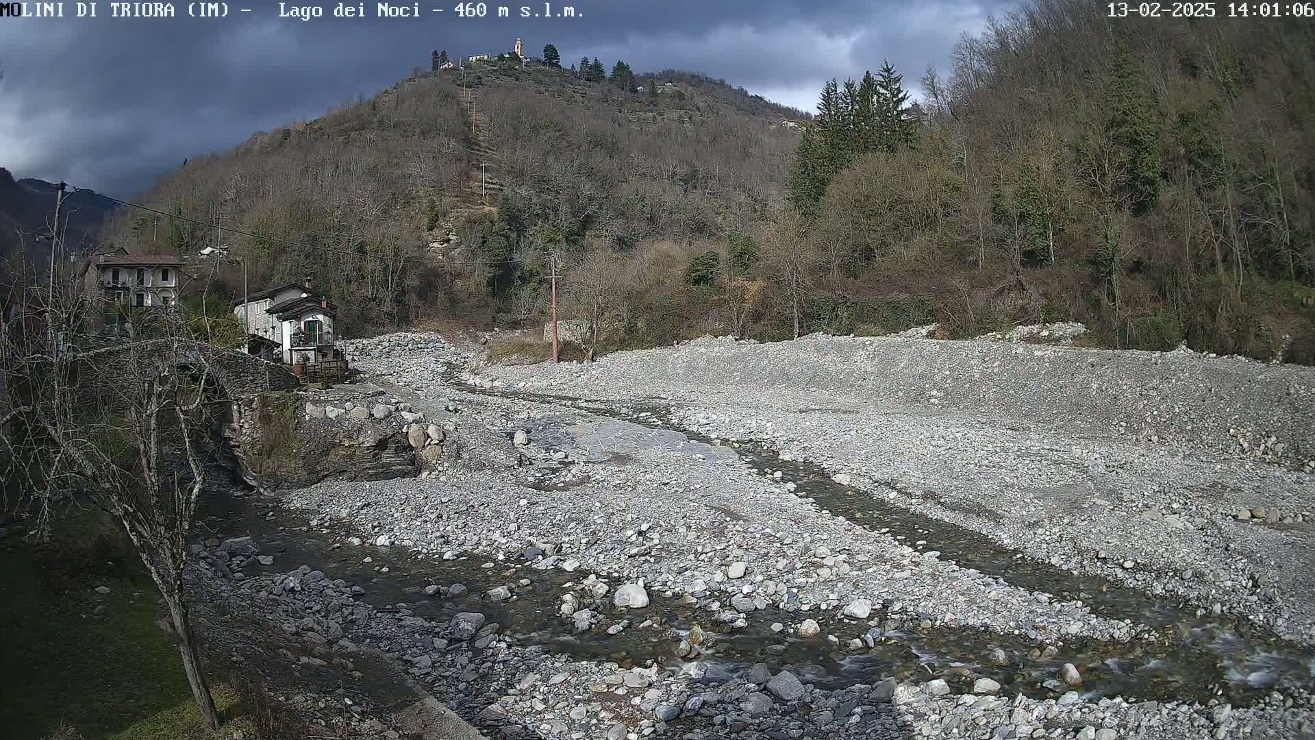 A rocky, mostly dry riverbed with a small stream flows through it, nestled between a village and a hillside under a partly cloudy sky.