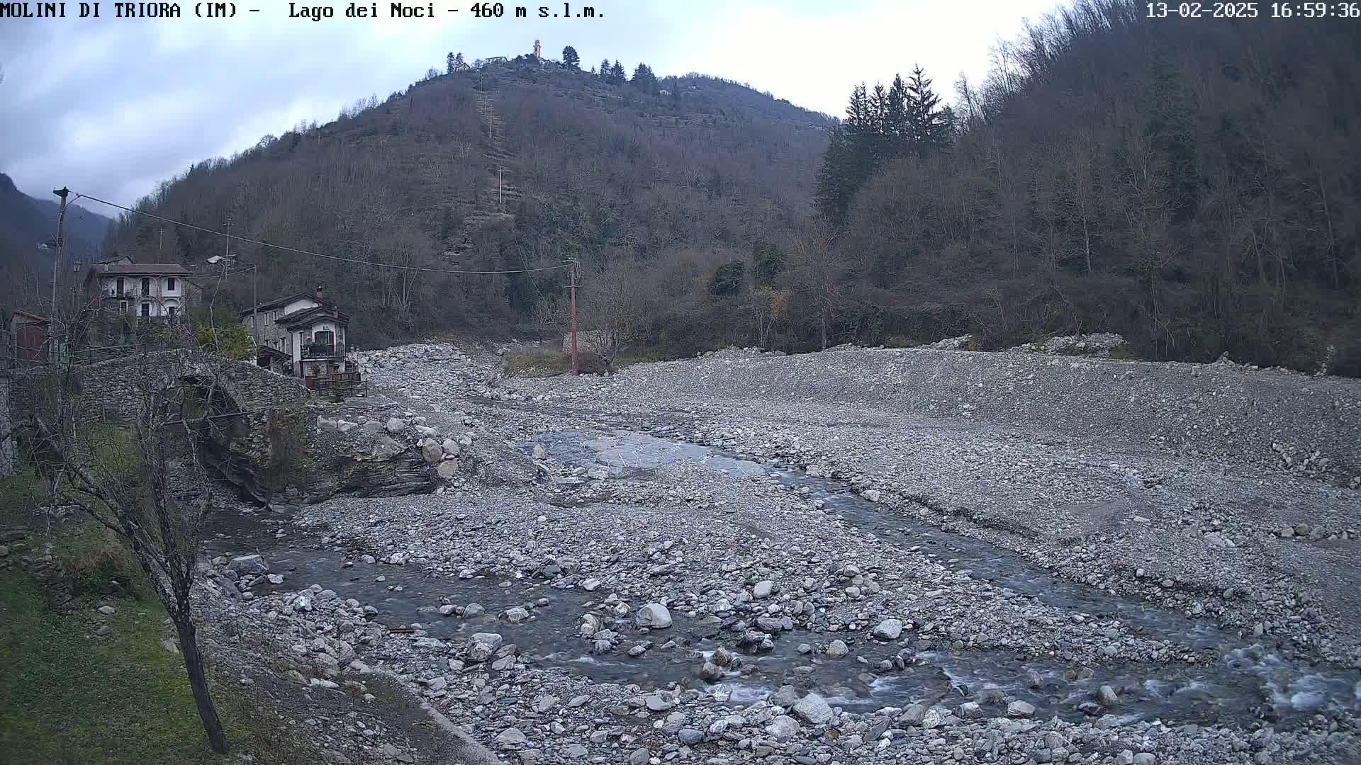 A rocky, mostly dry riverbed with a small stream flows through it, nestled between a village with stone buildings and a hillside covered in leafless trees under an overcast sky.