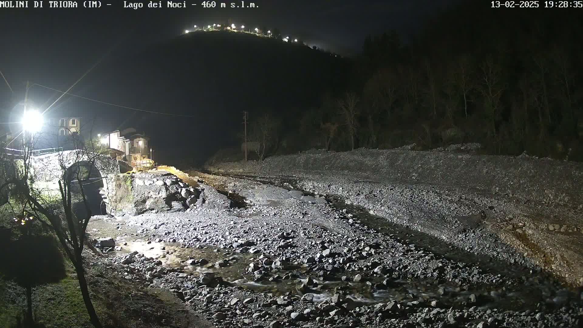 A rocky stream flows under a stone bridge at night, near a village nestled at the base of a hill.