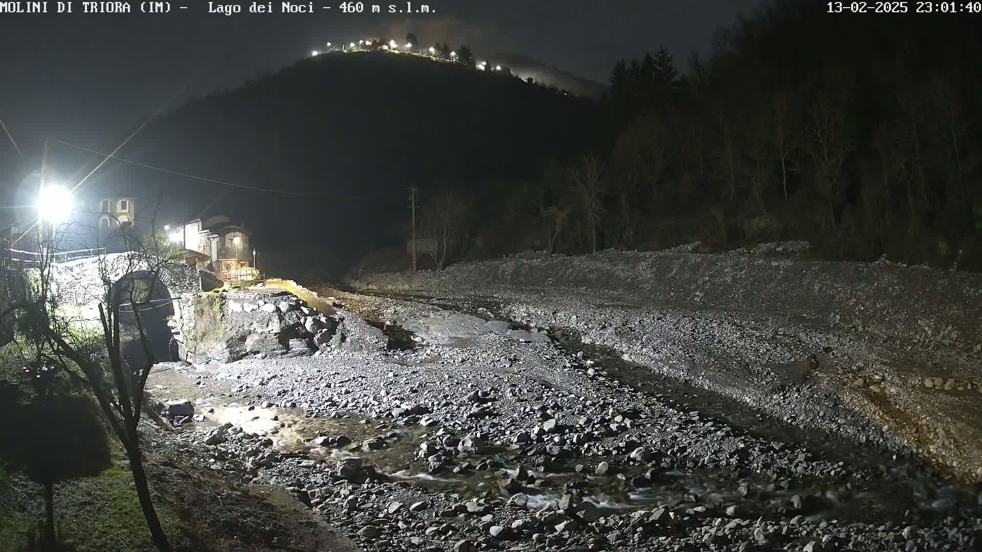 A rocky stream flows under a stone bridge at night, past a small village nestled at the base of a dark, hilltop.