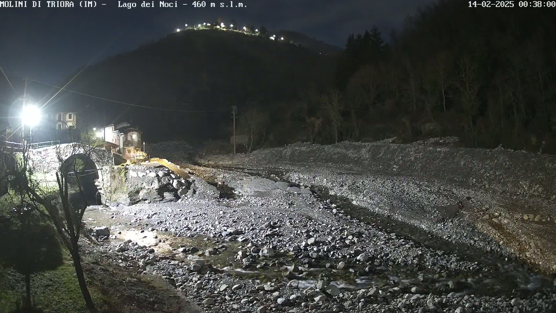 A nighttime view of a rocky riverbed with a small stream flowing through it, passing under a stone arch bridge near a small village nestled at the base of a hill.