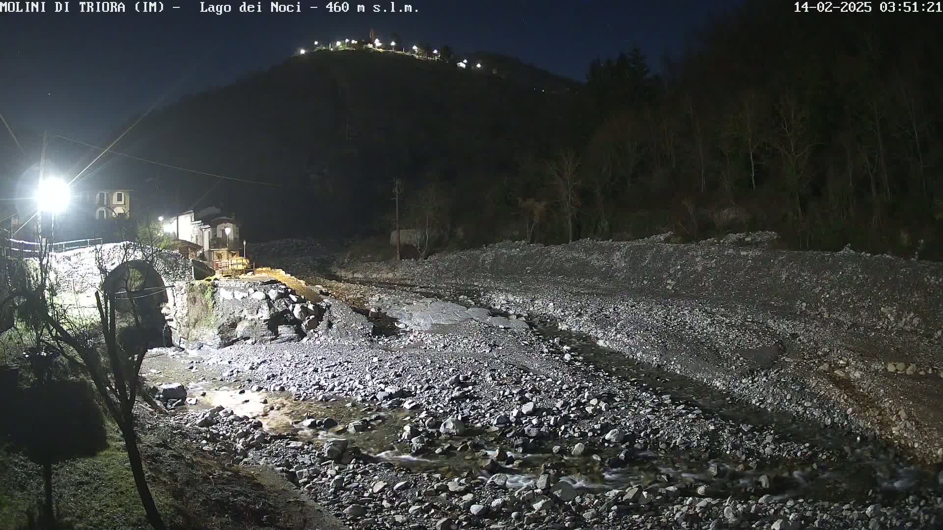 A stone bridge and buildings are illuminated at night beside a rocky stream under a clear, dark sky.