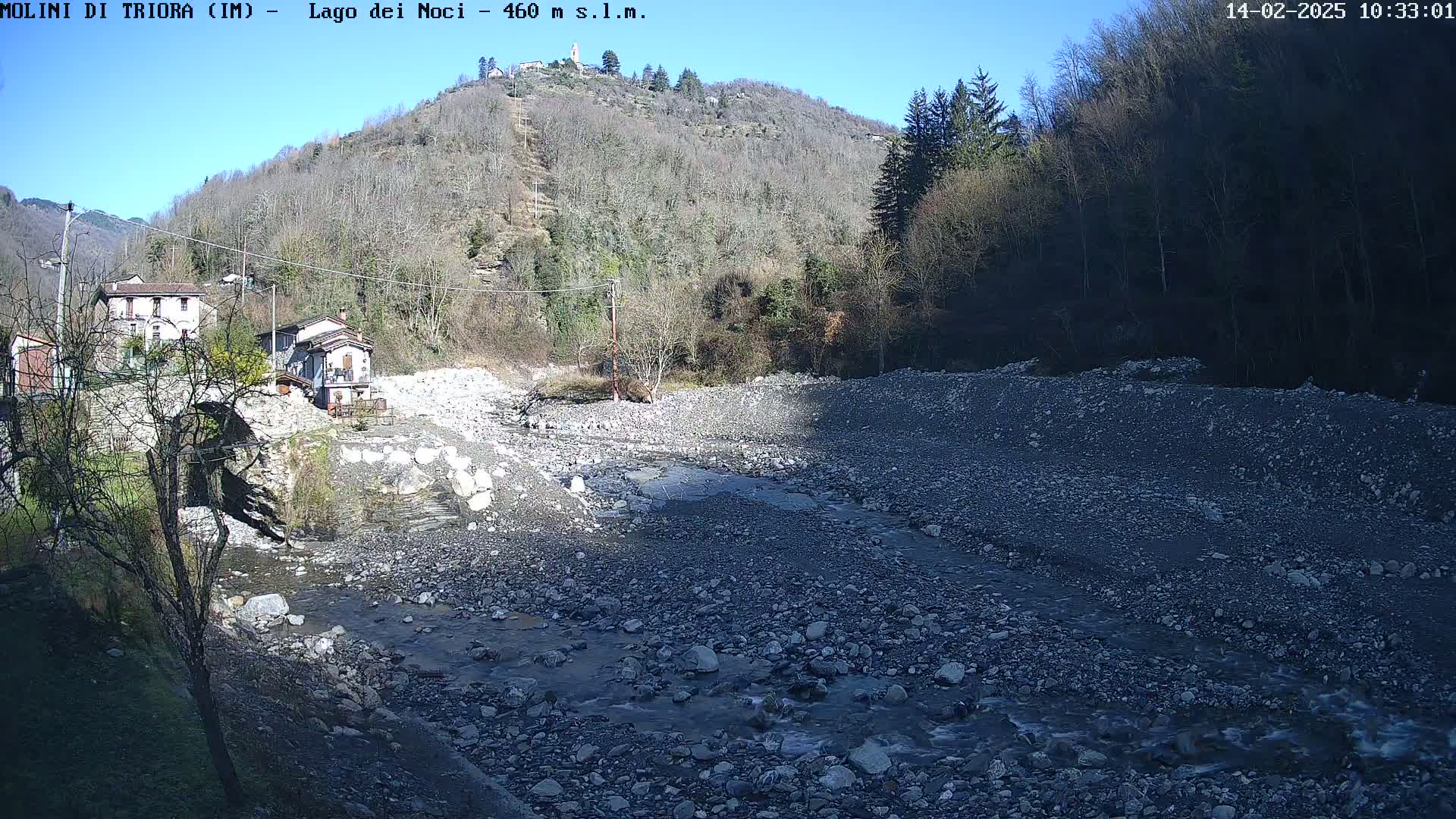 A rocky, shallow river flows past a village nestled at the base of a leafless, sunlit hill under a clear blue sky.