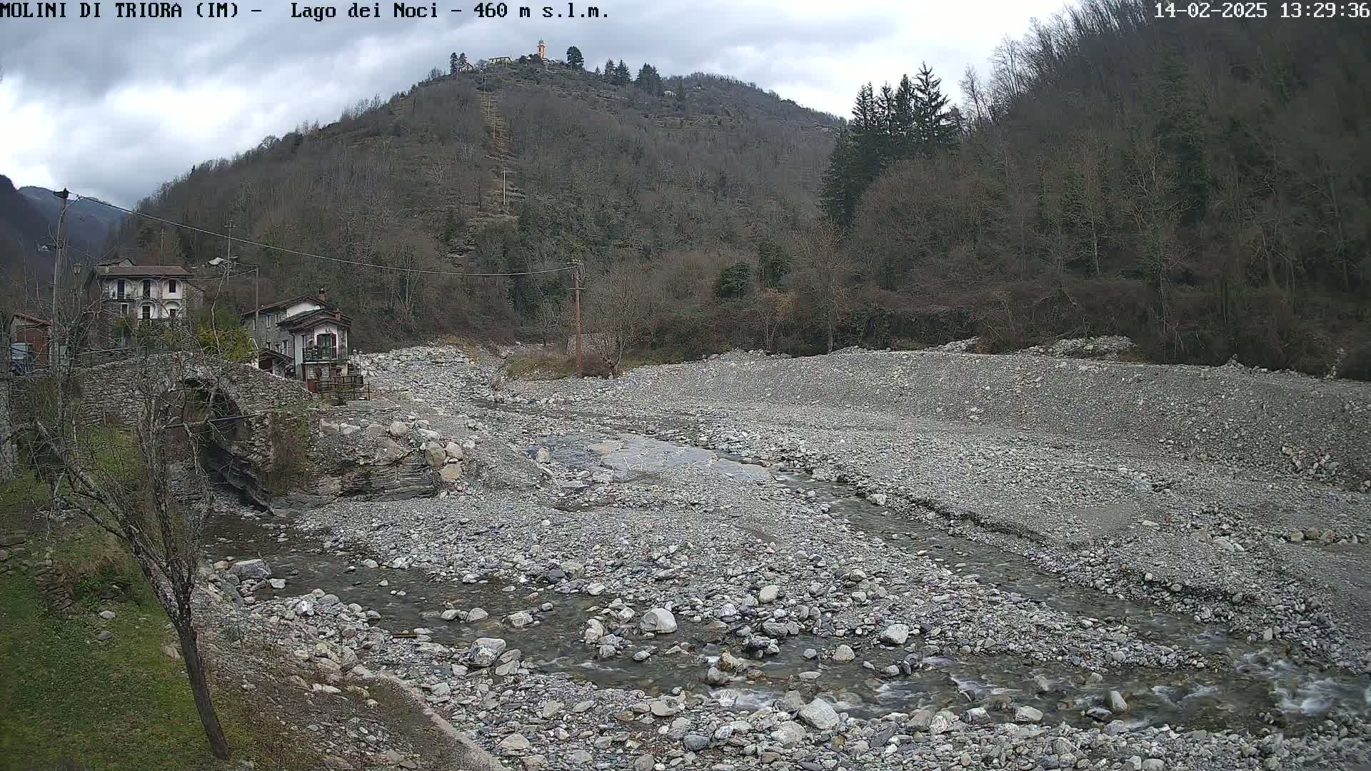 A rocky, mostly dry riverbed with a small stream flows through it, under a cloudy sky, near a stone bridge and houses nestled against a hillside of leafless trees.