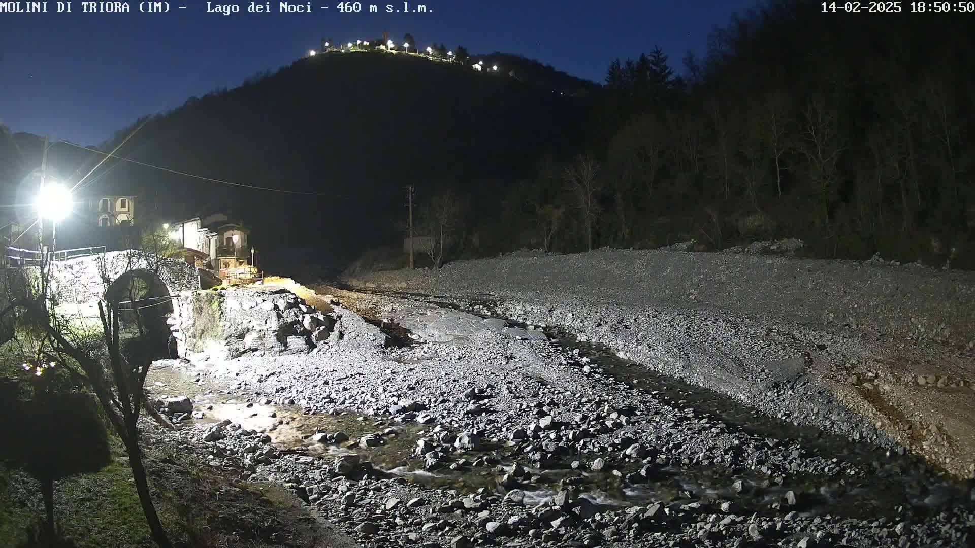 A nighttime view of a stone bridge over a shallow, rocky river, with a village and a hill illuminated by artificial lights in the background under a clear, dark sky.