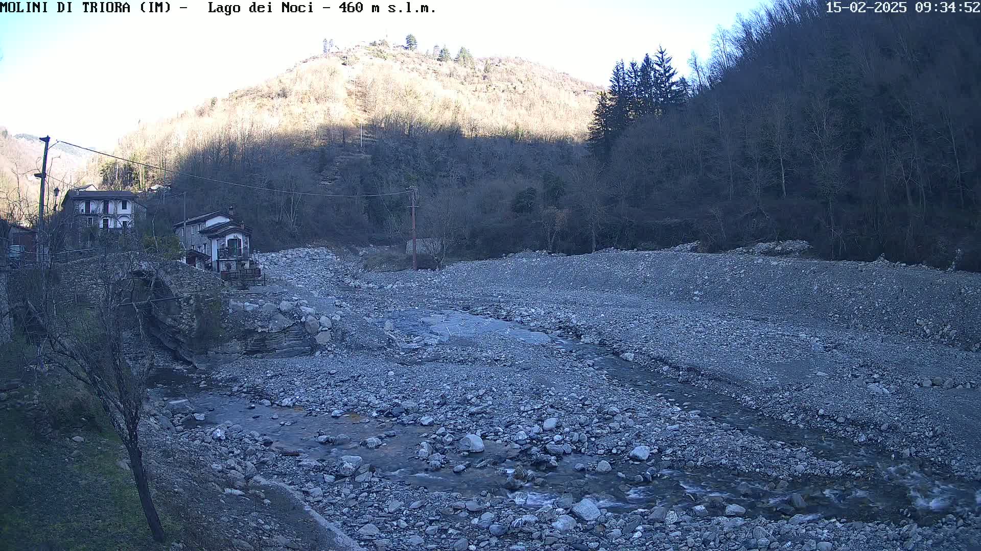 A rocky, dry riverbed with a small stream flows through it, near a village nestled in a valley under a clear sky.