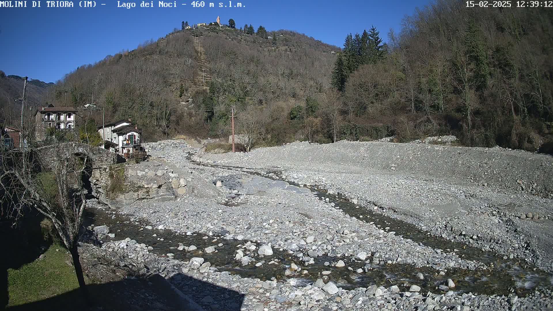 A rocky, dry riverbed with a small stream flows through it, nestled between a village and a hill covered in leafless trees under a clear, sunny sky.