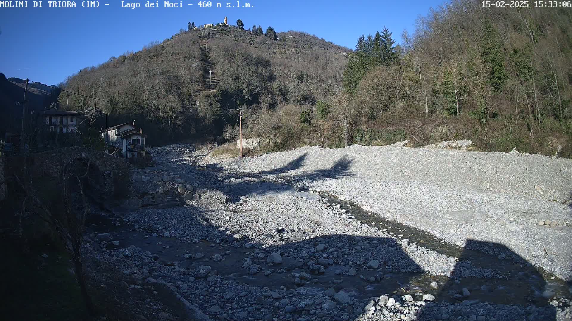 A rocky, shallow riverbed with a small stream of water flows through it, under a clear, sunny sky, near a village nestled against a hillside covered in leafless trees.