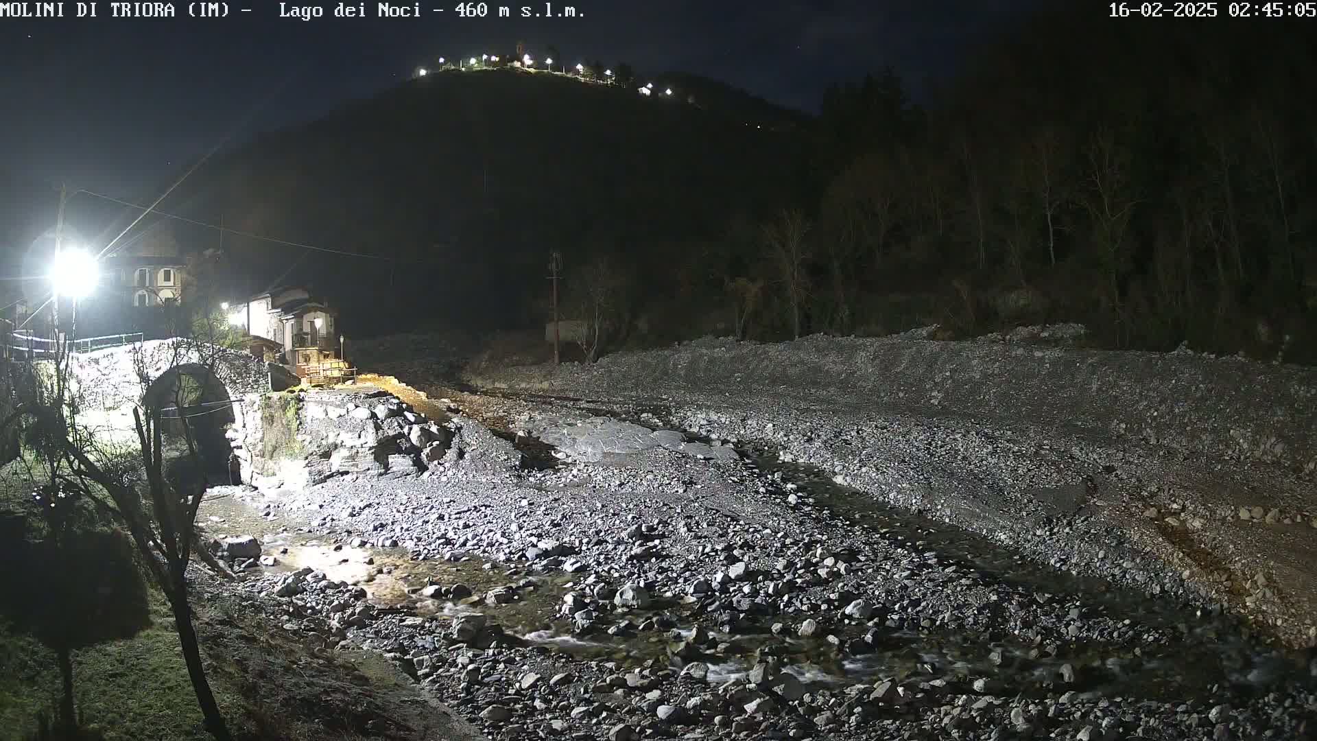 A rocky, shallow stream flows under a stone bridge at night, past buildings illuminated by artificial light against a dark hillside.