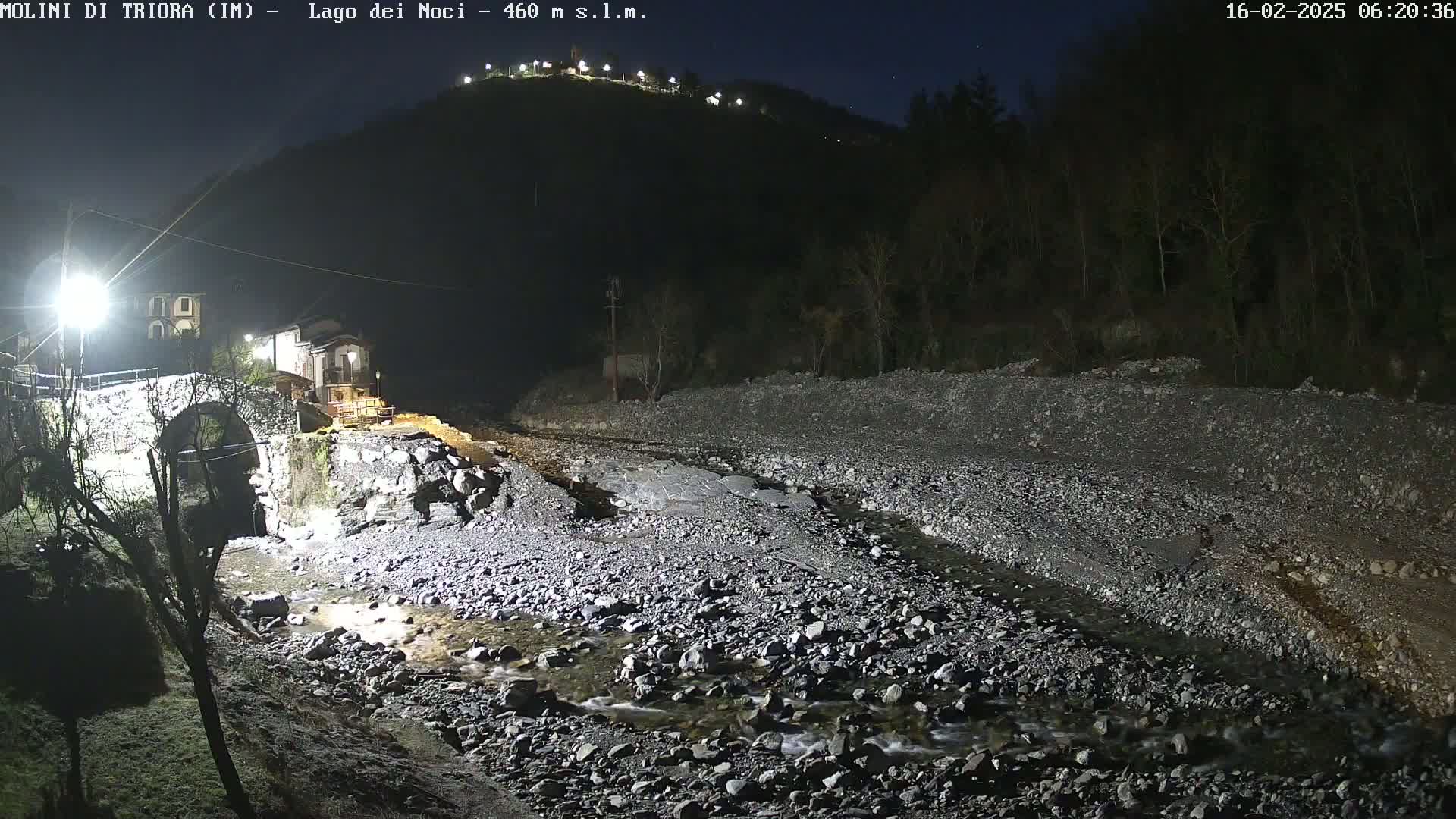 A rocky, shallow stream flows under a stone arch bridge at night, lit by artificial light, with a dark hillside rising in the background under a clear, starlit sky.