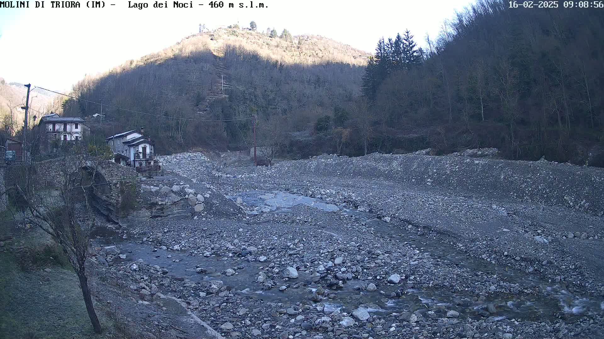 A rocky, mostly dry riverbed with a small stream flows through it, near two buildings and a stone bridge, under a clear, sunny sky;  hills covered in leafless trees rise in the background.