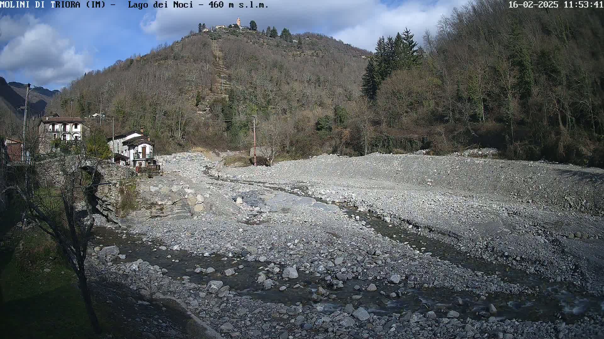 A mostly sunny day reveals a rocky, dry riverbed with a small stream flowing through it, near a village nestled against a hillside of bare, winter trees.