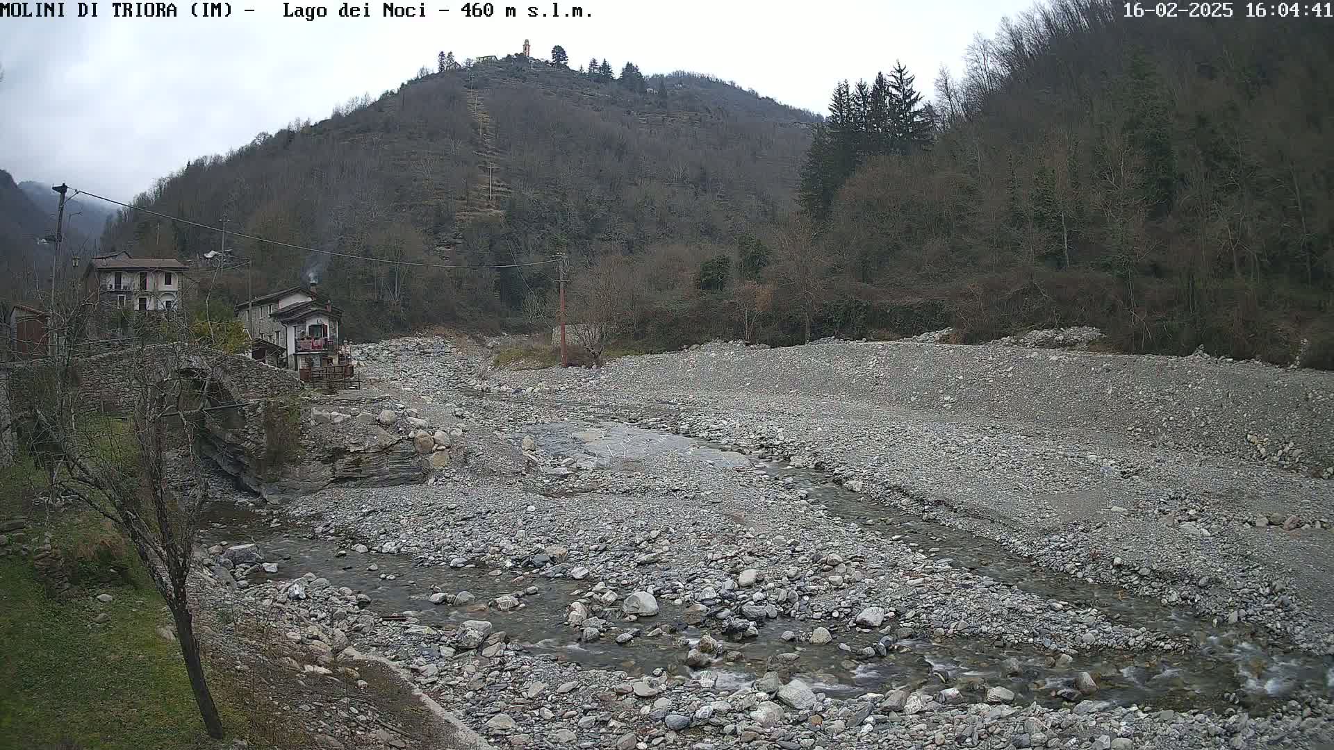 A rocky, mostly dry riverbed with a thin stream of water flows through it, past a stone bridge and houses nestled against a hillside under an overcast sky.