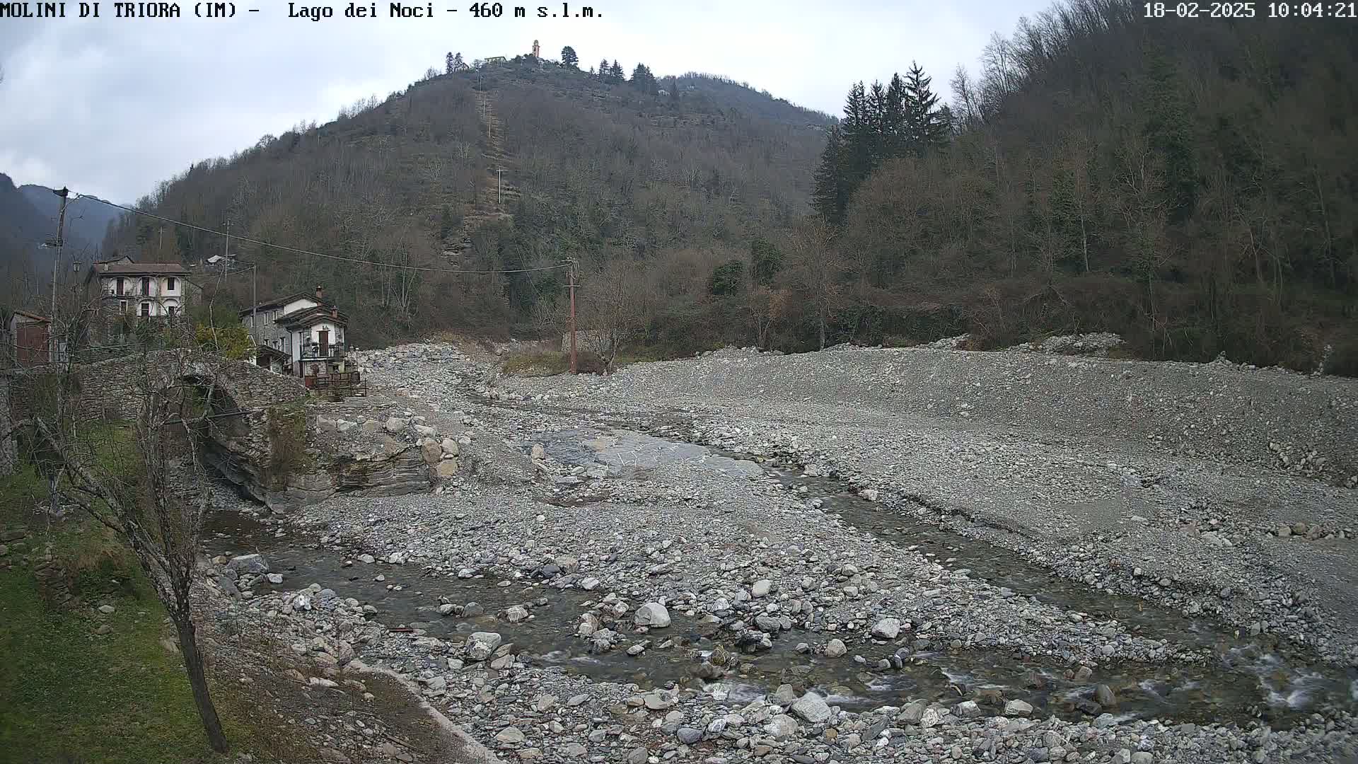 A rocky, dry riverbed with a small stream flows through it, near a stone bridge and buildings nestled at the base of a wooded hillside under an overcast sky.