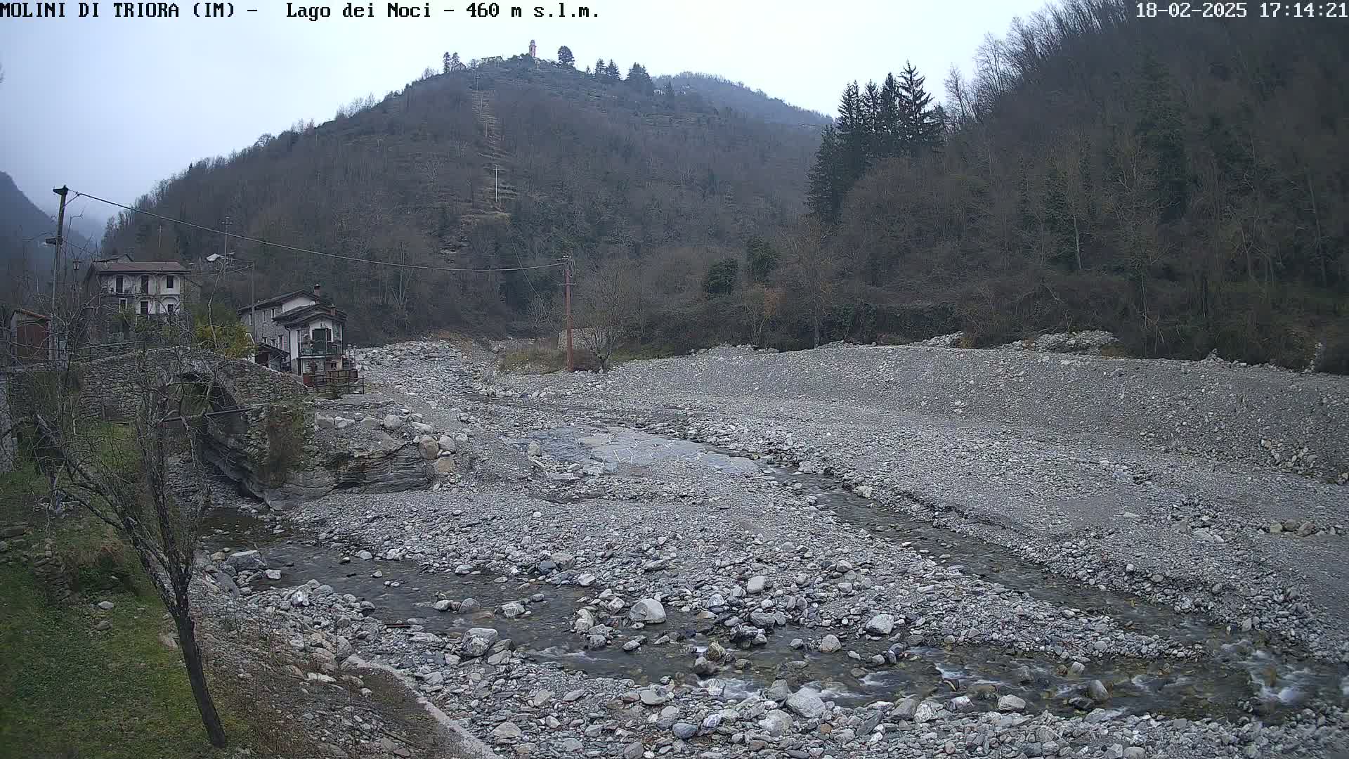 A rocky riverbed with a small stream flows through it, past a village nestled against a hillside under an overcast sky.