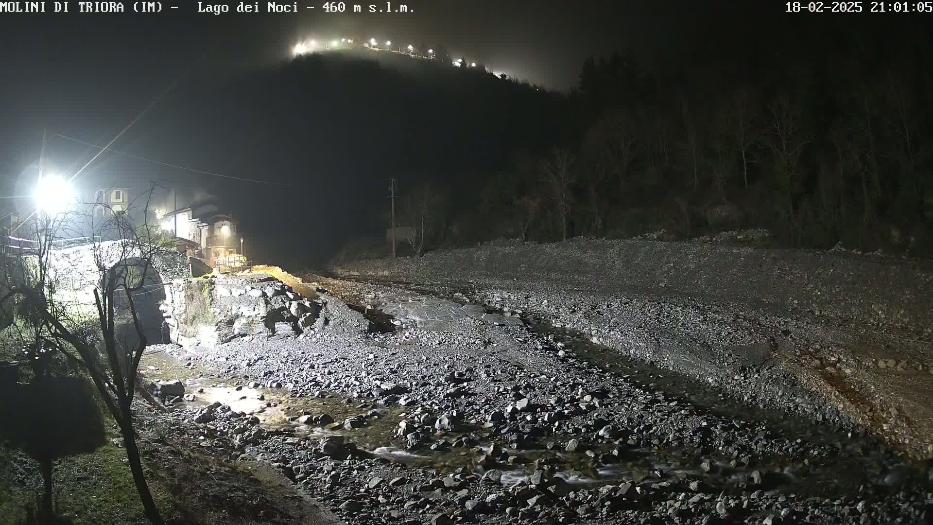 A nighttime, foggy view of a rocky stream bed illuminated by artificial light, with a stone bridge and building visible in the foreground and a hilltop in the background.