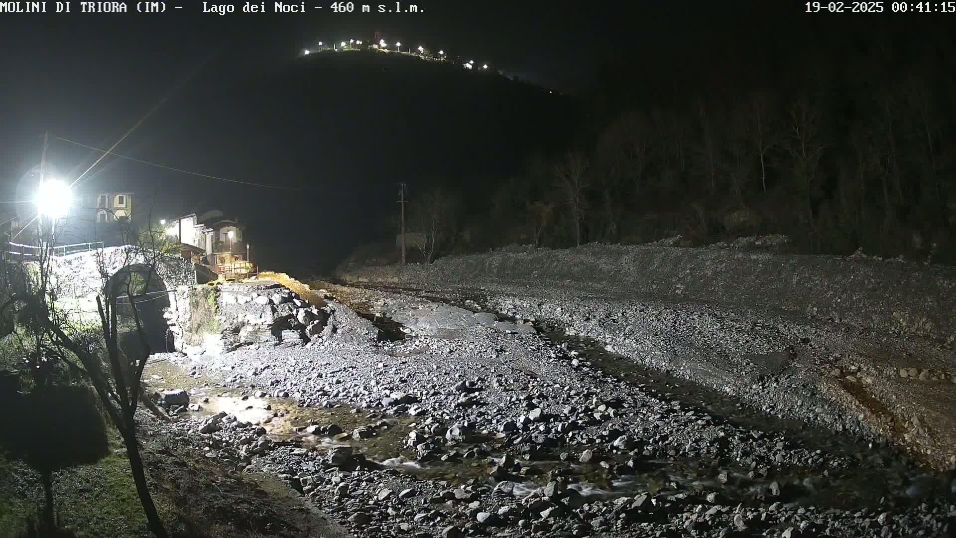 A nighttime view of a rocky stream bed with a small stream flowing through it, a stone bridge, and buildings illuminated on a hill under a clear, dark sky.