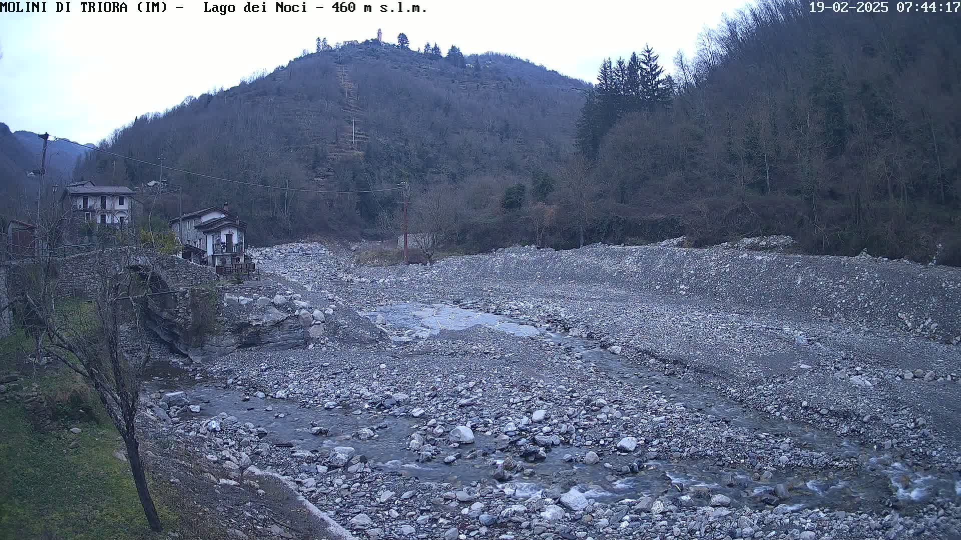 A rocky, shallow river flows through a wide, dry riverbed past a couple of houses nestled at the base of a tree-covered hill under an overcast sky.
