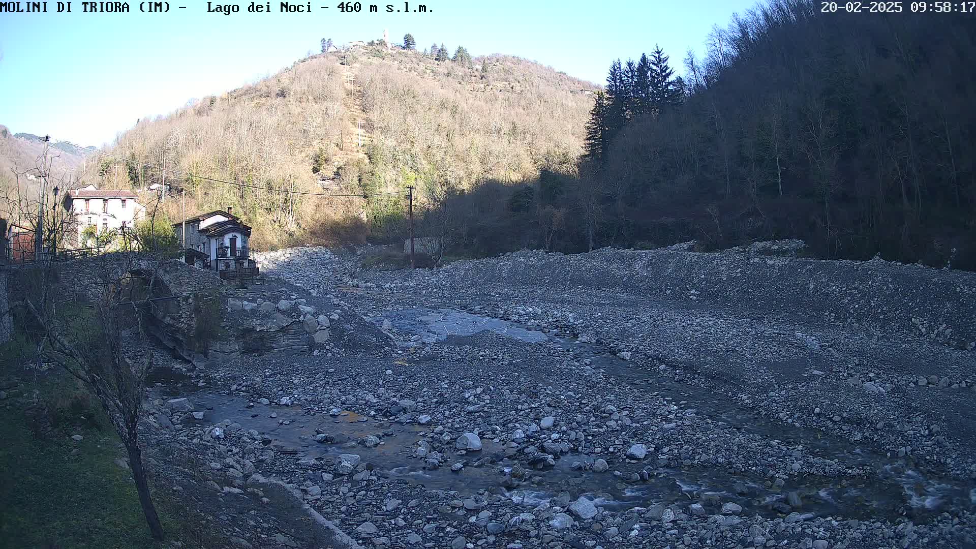 A rocky, dry riverbed with a small stream flows through it, near two buildings and a stone bridge, under a clear, sunny sky, with a hillside covered in leafless trees in the background.