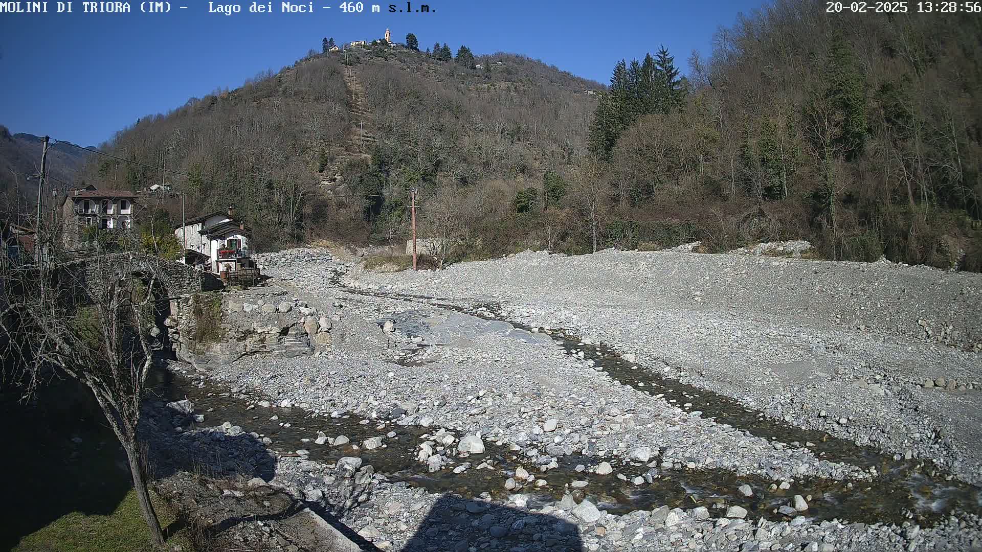A rocky, dry riverbed with a small stream running through it is visible below a hillside covered in leafless trees under a clear, sunny sky.
