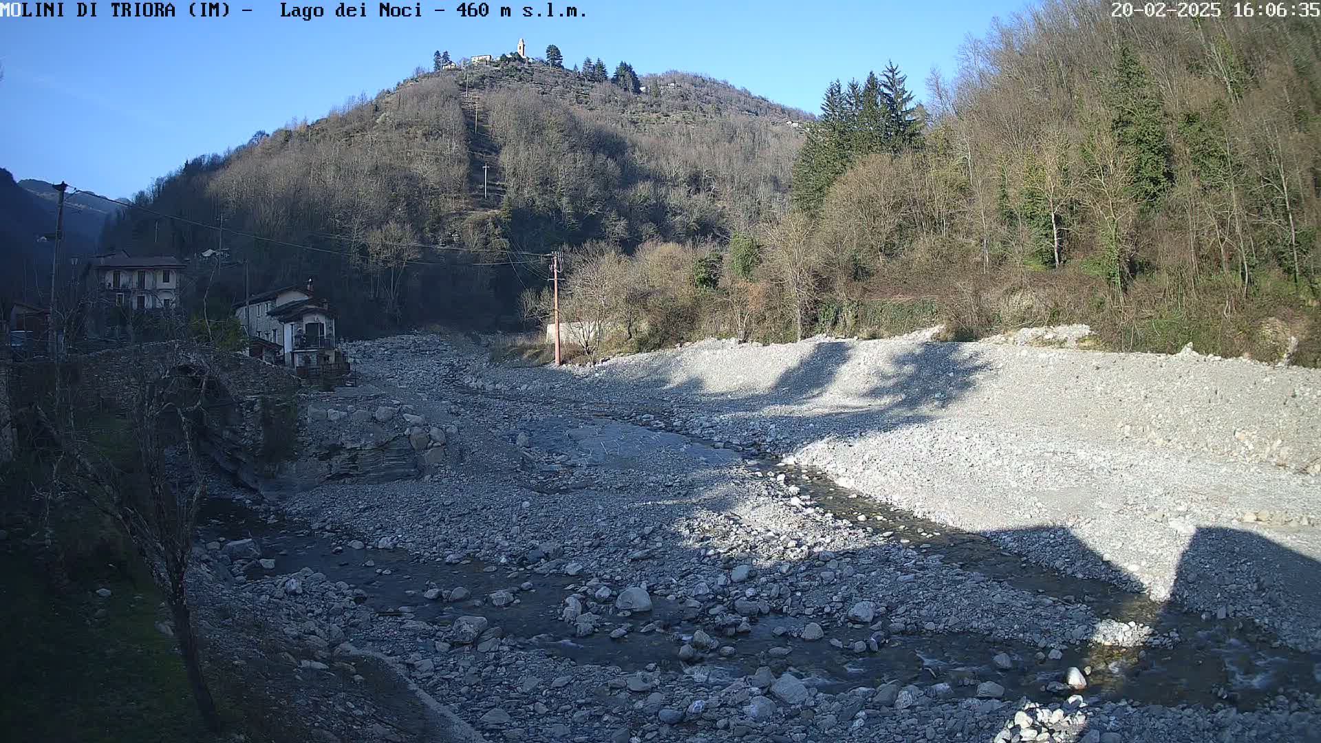 A rocky, shallow river flows through a valley under a clear blue sky, past buildings nestled at the base of a sparsely wooded hill.