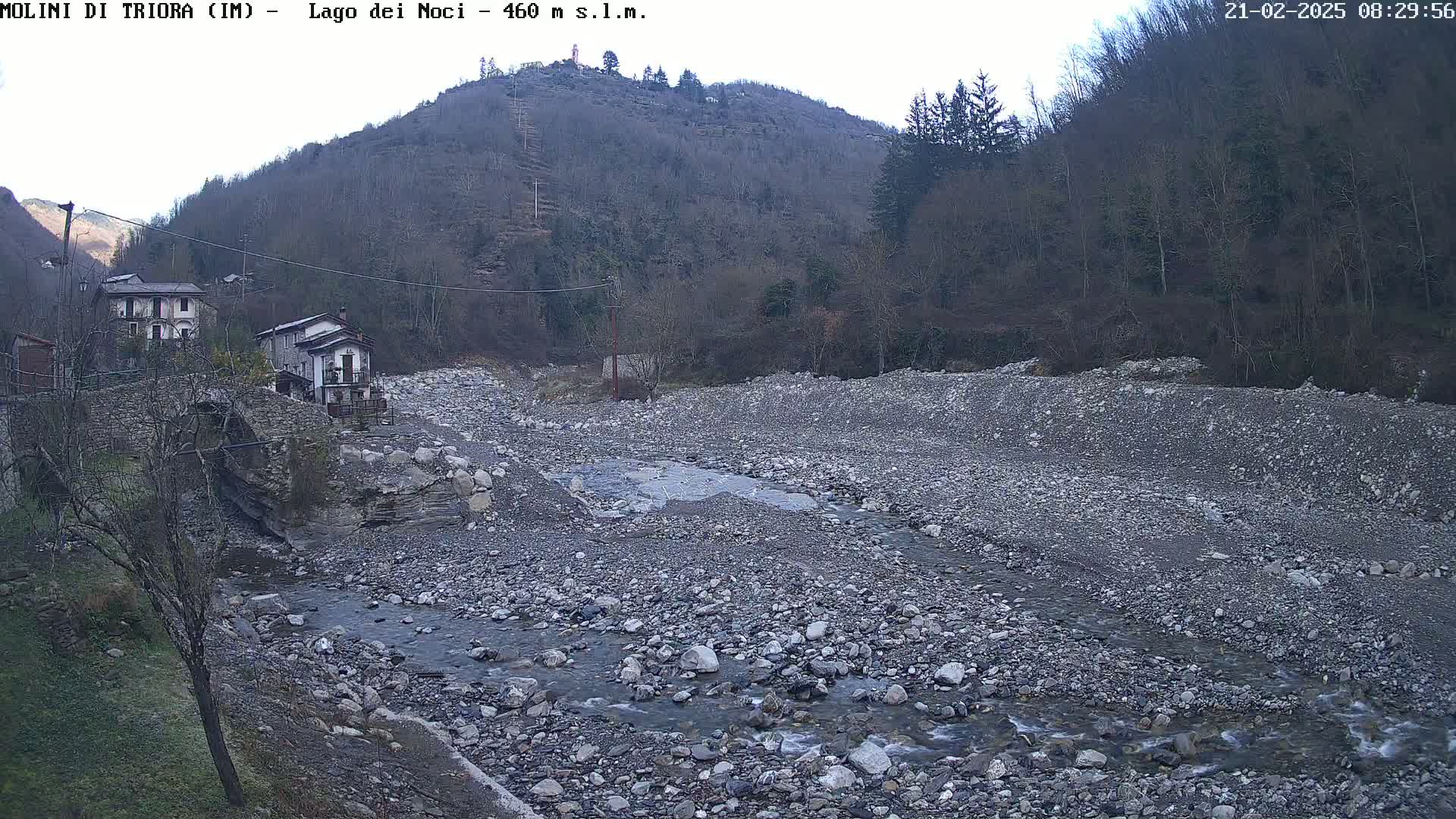 A rocky, dry riverbed with a small stream flows through it, situated below a hillside covered in leafless trees under a mostly clear sky.