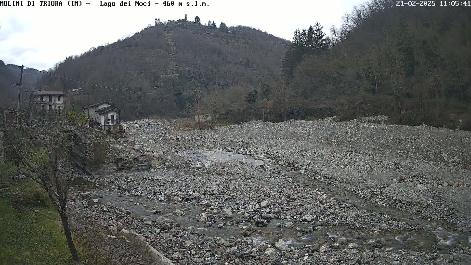 A rocky, shallow stream flows through a mostly dry riverbed near a small village nestled against a hillside under an overcast sky.