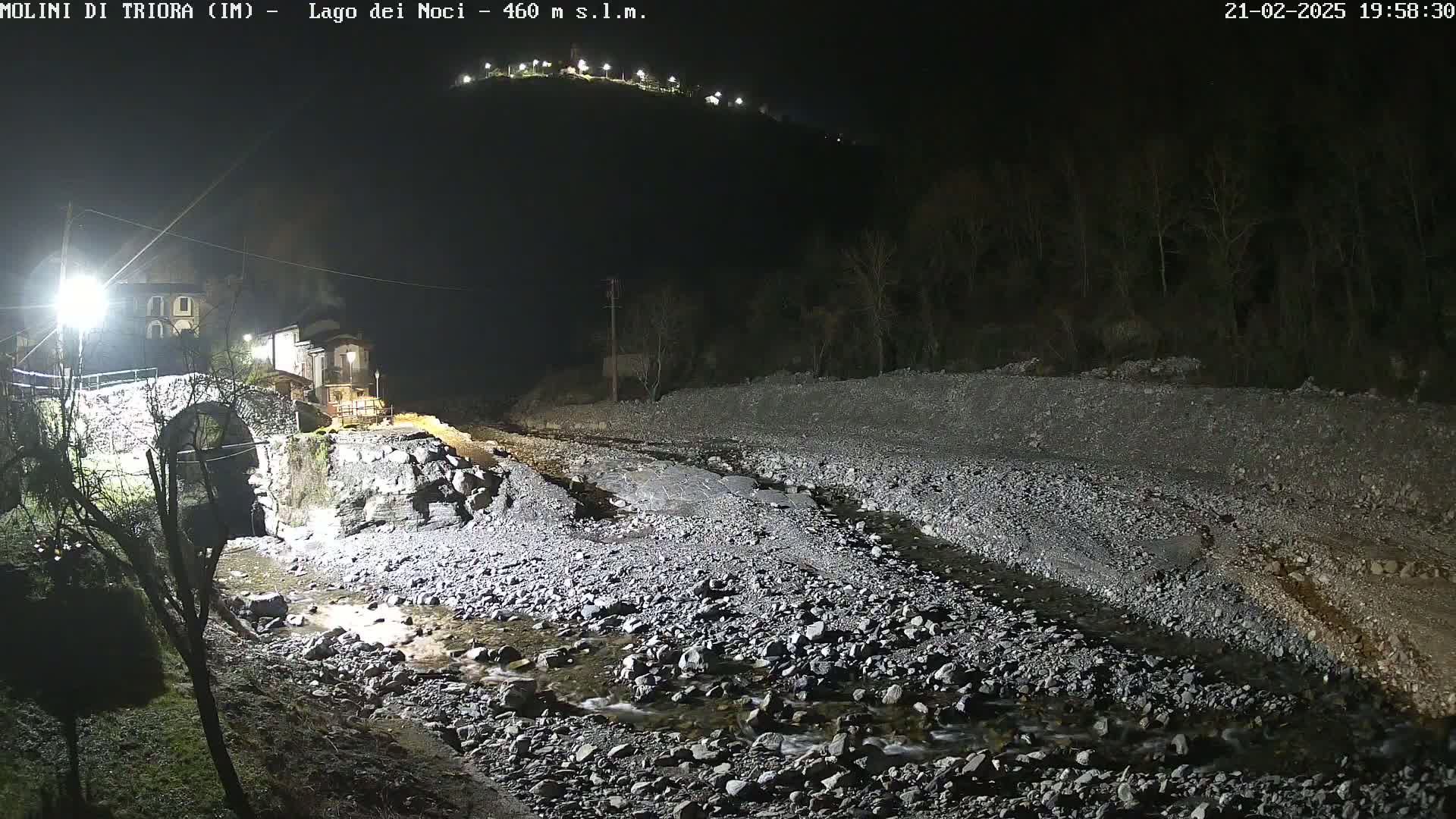 A nighttime view of a rocky stream bed illuminated by artificial lights, with a stone bridge and buildings visible in the background.