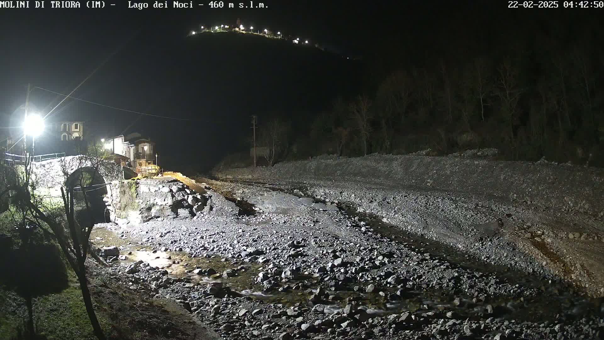 A nighttime view of a rocky riverbed with a stone bridge and buildings visible under artificial light.