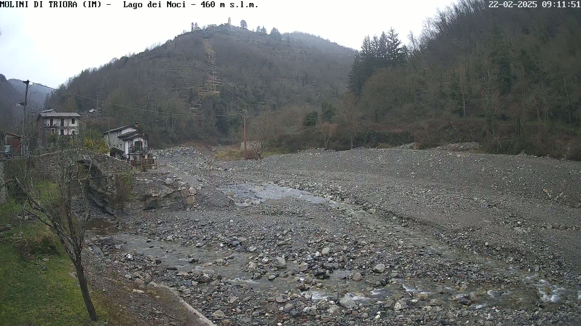 A rocky, dry riverbed with a small stream flows through it, past a stone bridge and houses nestled against a hillside under an overcast sky.