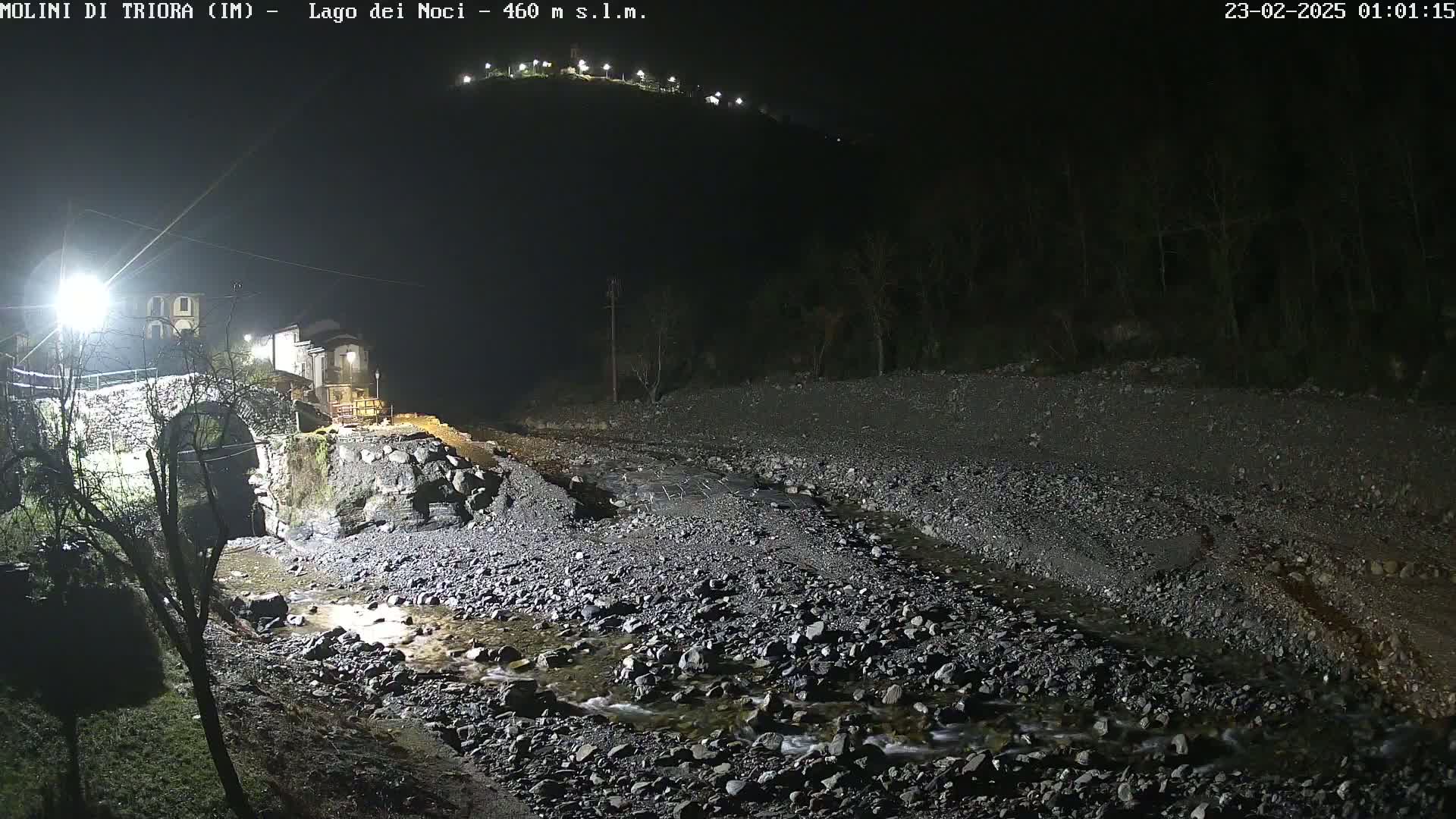 A nighttime view of a rocky streambed with a stone bridge and buildings in the background under artificial light.