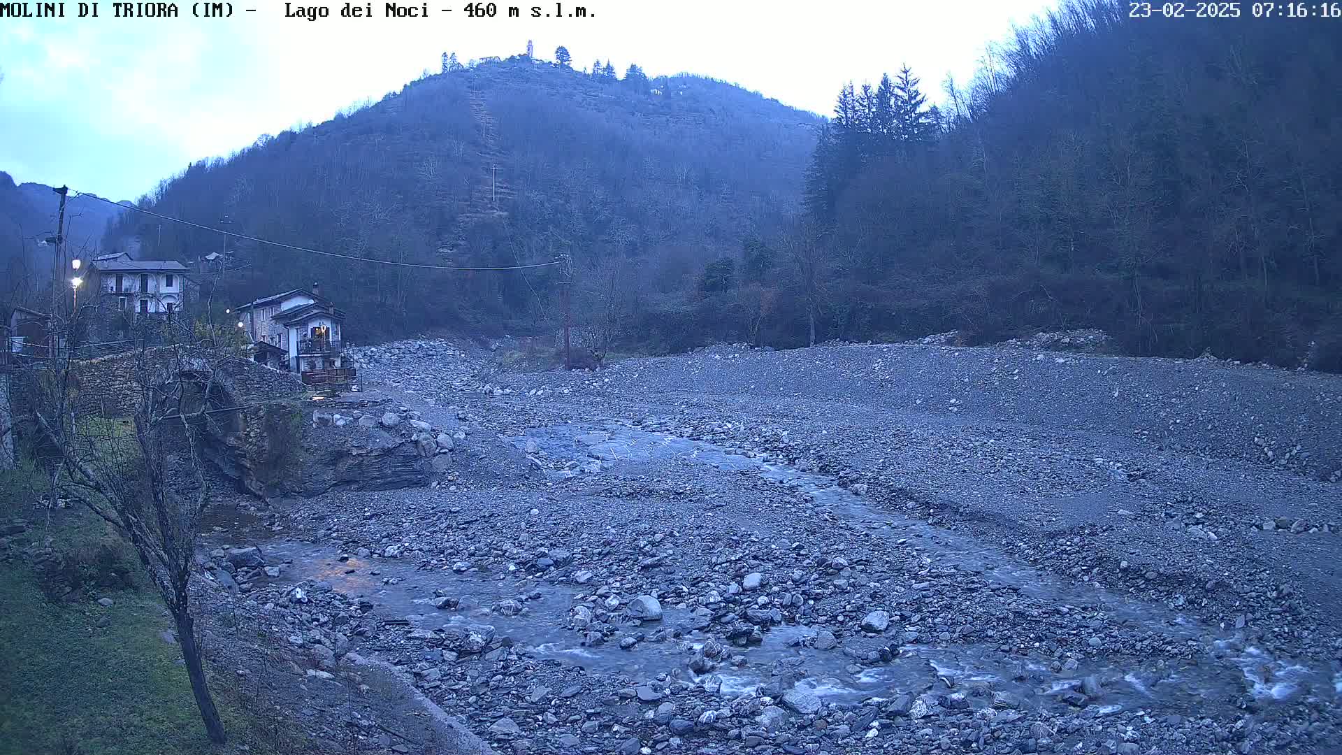 A rocky, dry riverbed with a small stream flows through it, nestled between a village and a hillside under a dusky sky.